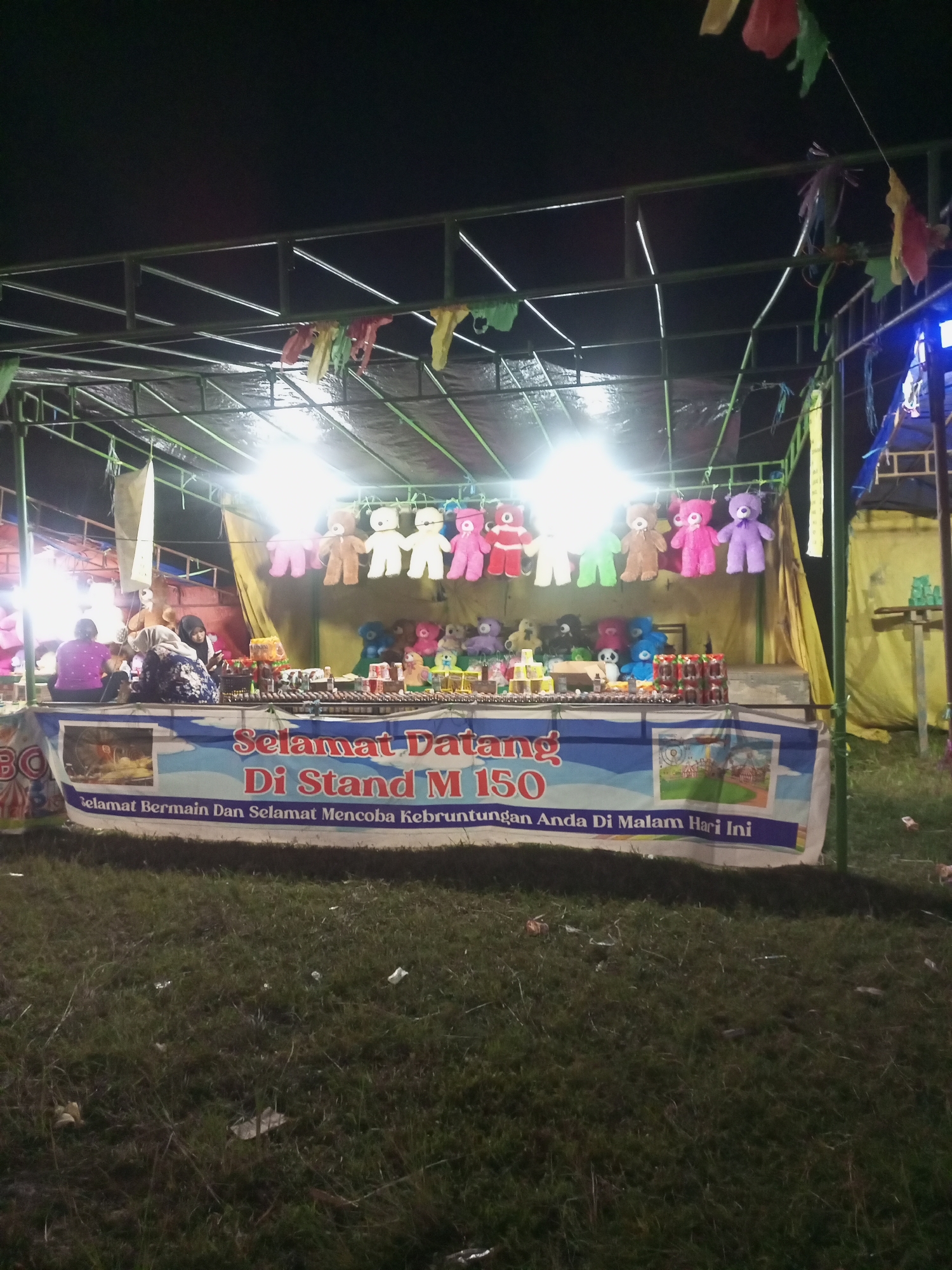 This image shows a nighttime scene at an outdoor stall, likely part of a fair or carnival. The stall is illuminated by bright lights, casting a glow on rows of colorful stuffed animals, primarily teddy bears, hung from the ceiling. Below the stuffed animals, a counter is laden with more toys, drinks in cans, and what appear to be small prize boxes or game elements. A banner across the front of the stall reads "SELAMAT DATANG DI STAND M 150 SELAMAT BERMAIN DAN SELAMAT MENCOBA KEBERUNTUNGAN ANDA DI MALAM HARI INI," which translates to "WELCOME TO STAND M 150, HAVE FUN AND GOOD LUCK TONIGHT." Two women are seated behind the counter, interacting with each other, with one woman wearing a purple shirt and the other a headscarf and patterned clothing. The stall is set up on a grassy area, with discarded debris visible on the ground. In the background, a yellow tarp and blue tent-like structure are visible, suggesting a larger event is taking place. The overall atmosphere is festive and commercial, with the focus on games and prizes.
