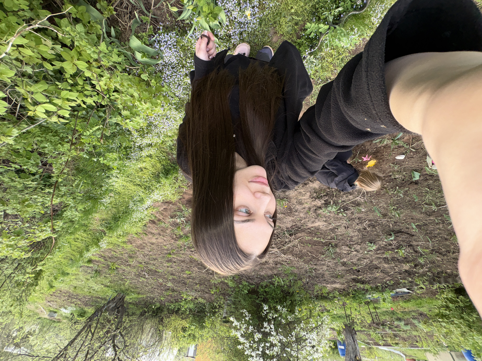 A selfie taken in an outdoor setting, possibly a backyard or garden, in Chaikovsky, Russia, shows a woman with long, brown hair and light eyes. She wears a black fleece jacket or coat. She stands in an area with mixed vegetation, including grass, flowering plants, and trees. The ground is partially covered in exposed soil. In the background, a residential area may be inferred with a glimpse of houses.