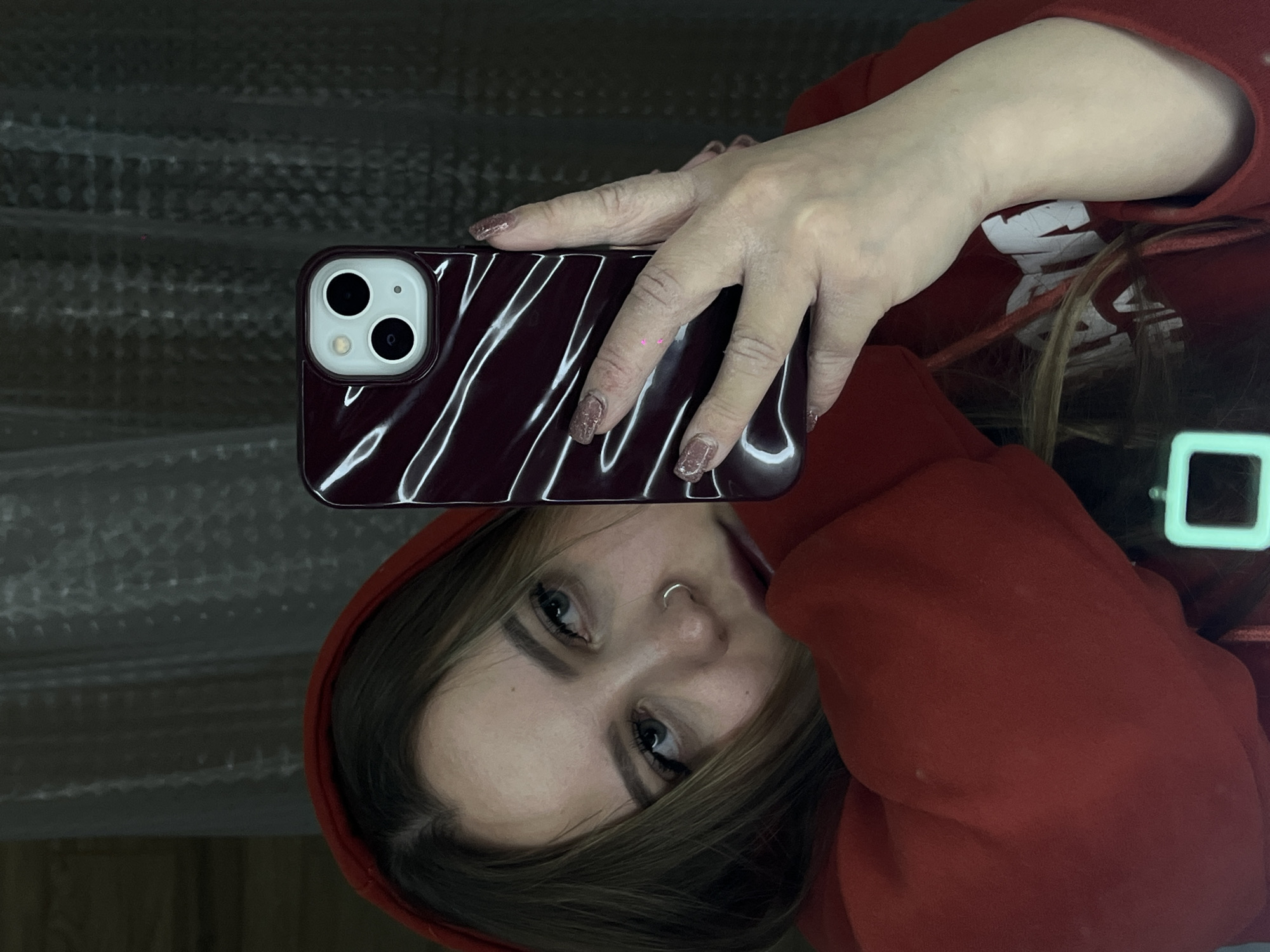 A young person, likely female, is captured in a reflective selfie, holding a smartphone. She is wearing a red hooded sweatshirt with the hood up, framing her face and dark hair. Her gaze is direct, and she has dark eyebrows and a silver hoop nose ring. Her right hand, adorned with glittery, brownish-pink nail polish, holds a smartphone. The phone appears to be an iPhone, housed in a glossy, dark red case with a prominent wavy or marbled pattern. The background behind her reflection is dark and textured, possibly a wall or bubble wrap, suggesting an indoor setting. No specific time of day or outdoor weather is discernible. Her expression is neutral. A subtle bright rectangular reflection is visible in the lower right of the frame. No legible text is visible in the image.