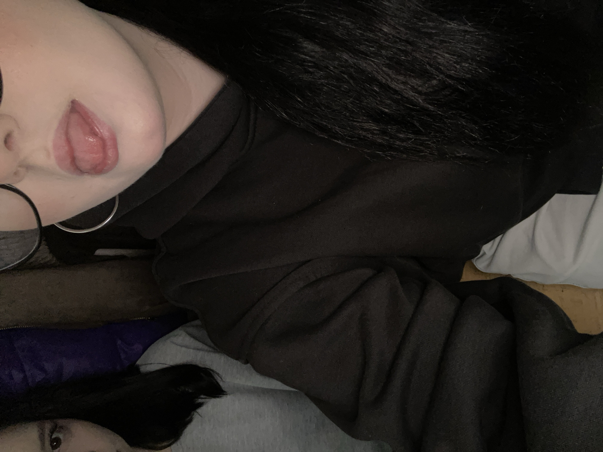 The image shows a person with black hair and glasses, wearing a black sweatshirt. The person's tongue is sticking out. The background includes a blue, gray, and brown surface. The location is not evident from the image.