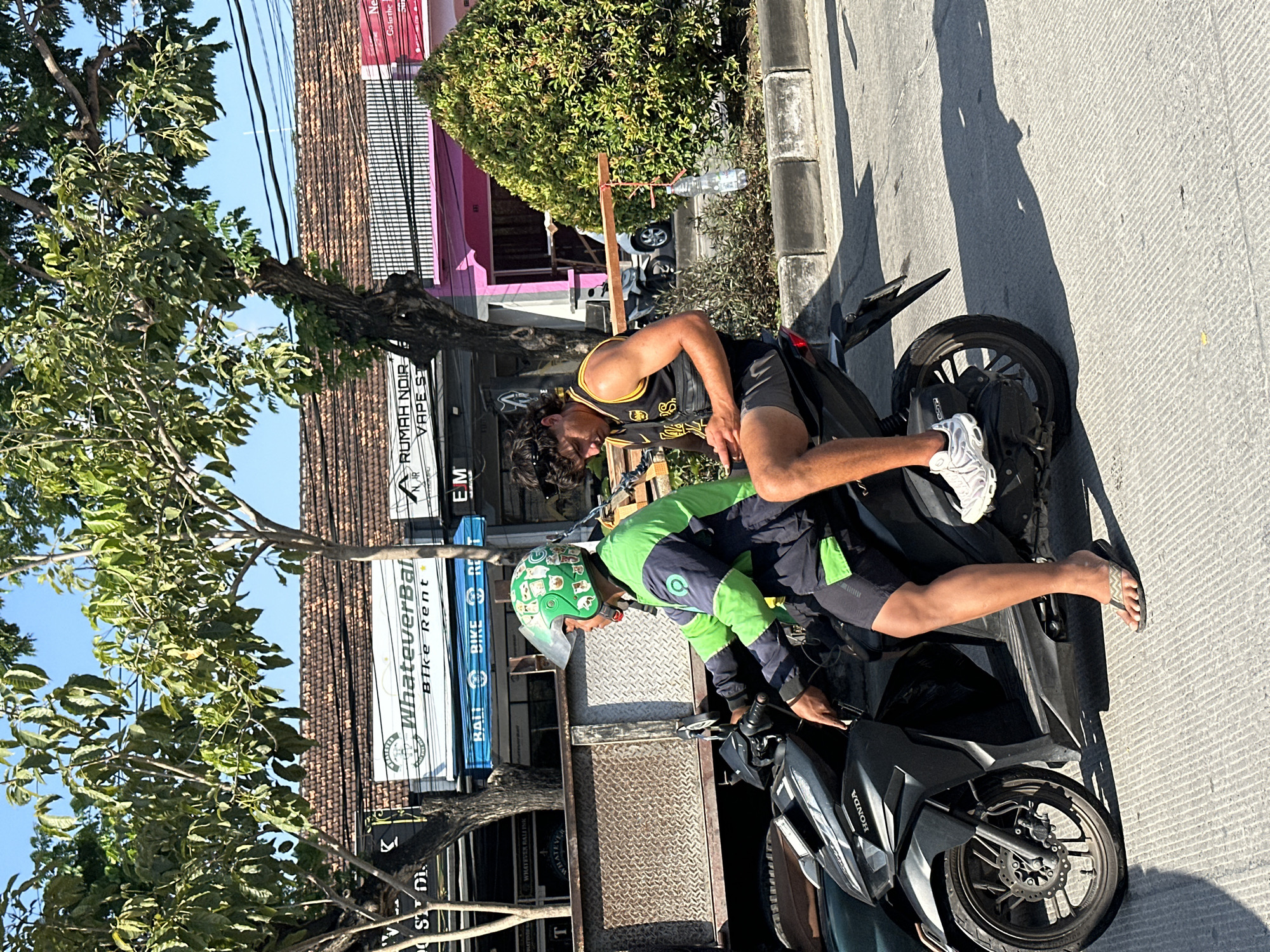 A medium shot captures two individuals on a dark gray Honda scooter on a street in Kuta, Indonesia, under a clear blue sky. The rider, wearing a green and dark blue jacket with a green helmet adorned with cartoon animal stickers, operates the scooter. The passenger, a young man with curly dark hair and sunglasses perched on his head, wears a black and yellow tank top and gray shorts. He is seated behind the rider, looking down at his mobile phone. The scooter has a license plate that is partially visible. In the background, buildings with tiled roofs and signs are visible, including "WhateverBal Bike Rent" and "RUMAH NOIR VAPE ST". Trees with lush green foliage line the street, casting shadows on the concrete road. A green bush is on the right side of the image, and a clear plastic bottle hangs from a wooden structure in front of it. Electrical wires are strung overhead. The scene depicts a typical street view in a bustling urban environment.