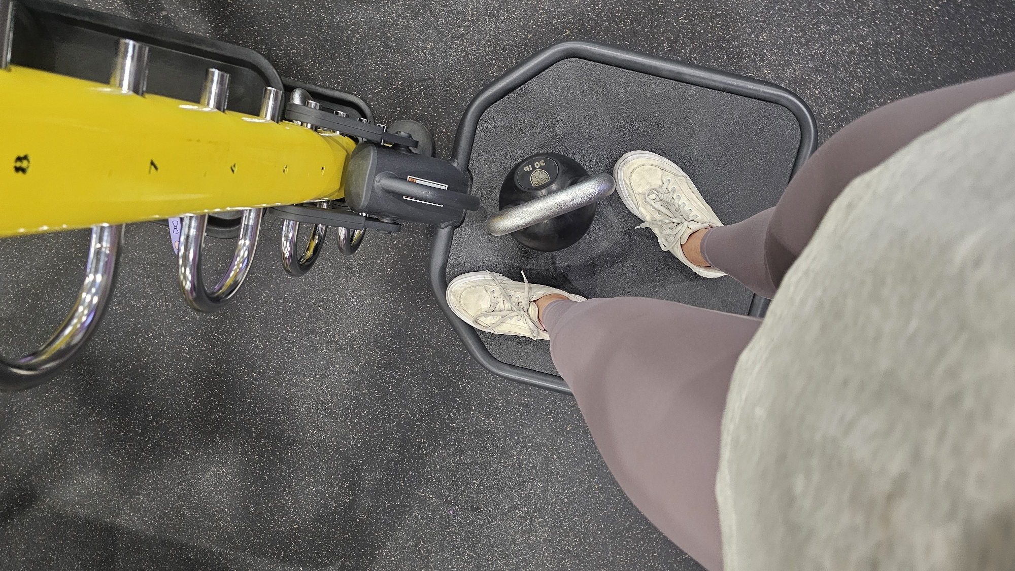The image presents a first-person, overhead view of an individual at a gym. The person is standing on a dark gray platform and is wearing mauve leggings and white tennis shoes with laces tied. A 30 lb kettlebell rests on the platform between their feet.
Adjacent to the platform is gym equipment with a yellow arm marked with numbered settings (likely to adjust the weight). The equipment is constructed with shiny, silver, curved rods and black components.
The gym floor visible around the equipment is a textured, dark gray surface. The image suggests an indoor environment, specifically within a fitness facility. There is no indication that this image was taken in Puebla, Mexico.