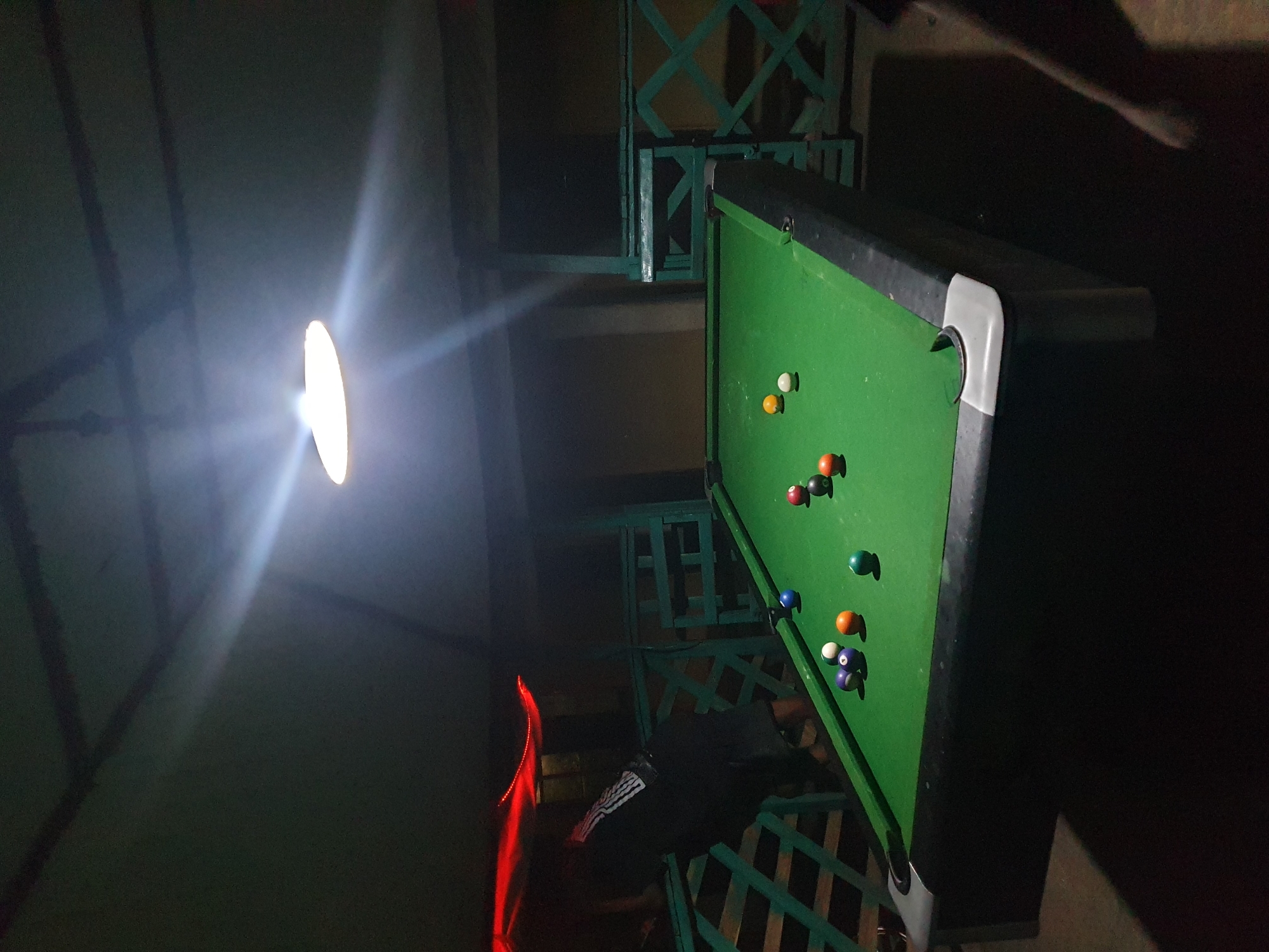 A pool table with multiple pool balls scattered on its green surface is under a bright light. The table is surrounded by a green, patterned railing, and a person in a dark shirt stands near the railing to the left of the table. The environment is dimly lit. The location is in Lokoja, Nigeria.