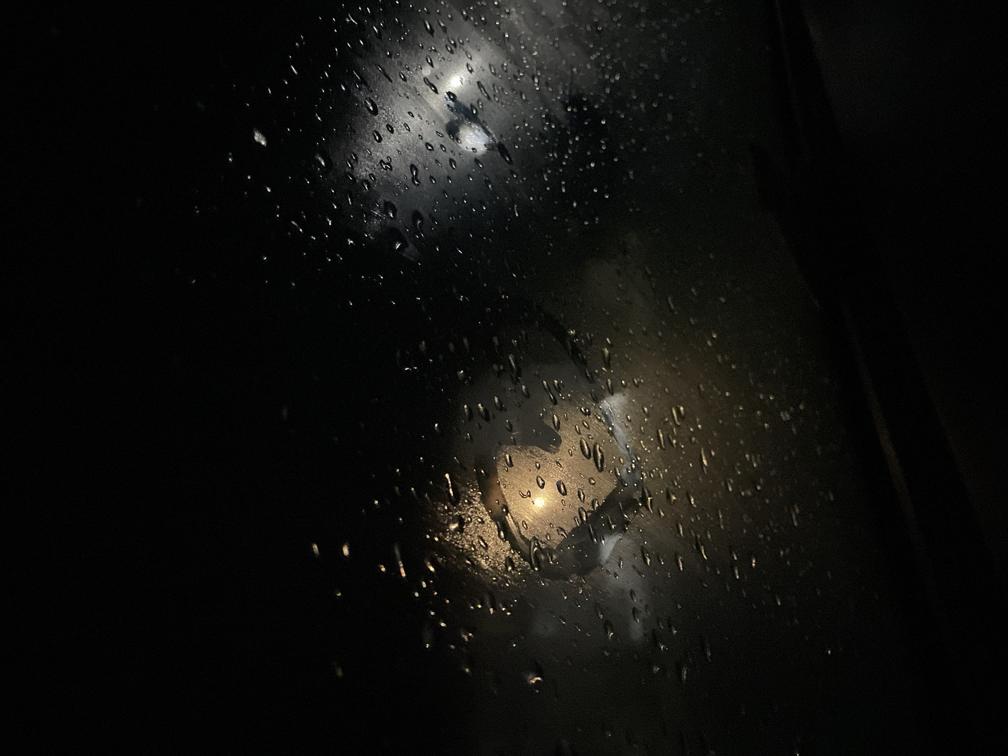 The image shows a window covered in raindrops at night. Through the wet glass, blurry lights are visible, likely from street lamps or vehicle headlights. The scene is very dark, with limited visibility beyond the rain-streaked window. There's no information available to identify if this photo was taken in Debelt, Bulgaria.