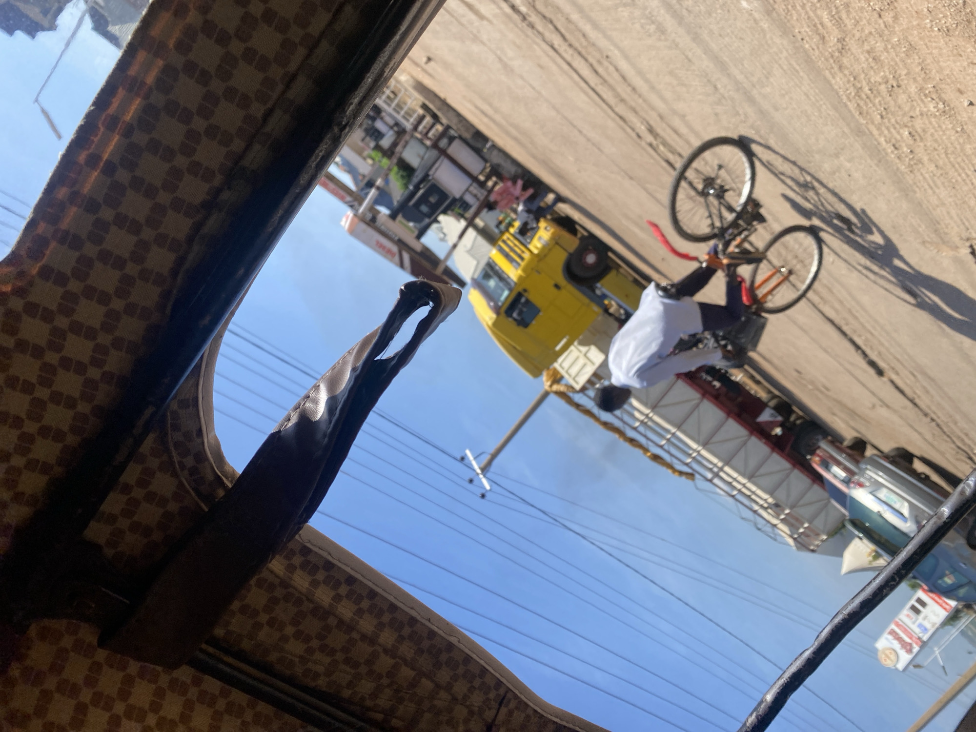 The image shows a daytime scene on a dusty road, possibly in Jos, Nigeria, viewed from the interior of a vehicle. In the foreground, the patterned fabric roof and a dark bar from the vehicle's interior frame are visible. The primary focus is a person riding a bicycle. The cyclist is wearing a white long-sleeved shirt and dark trousers, with their face obscured. They are on a red and black bicycle. Behind the cyclist, a large truck with a white cargo bed and a red frame is parked on the side of the road. Further in the background, a bright yellow truck is also visible, along with a structure that appears to be a gas station or a commercial building. Power lines are strung across the clear blue sky. The overall impression is of a busy street scene under a bright sun, with typical elements of urban Nigerian transportation and infrastructure.