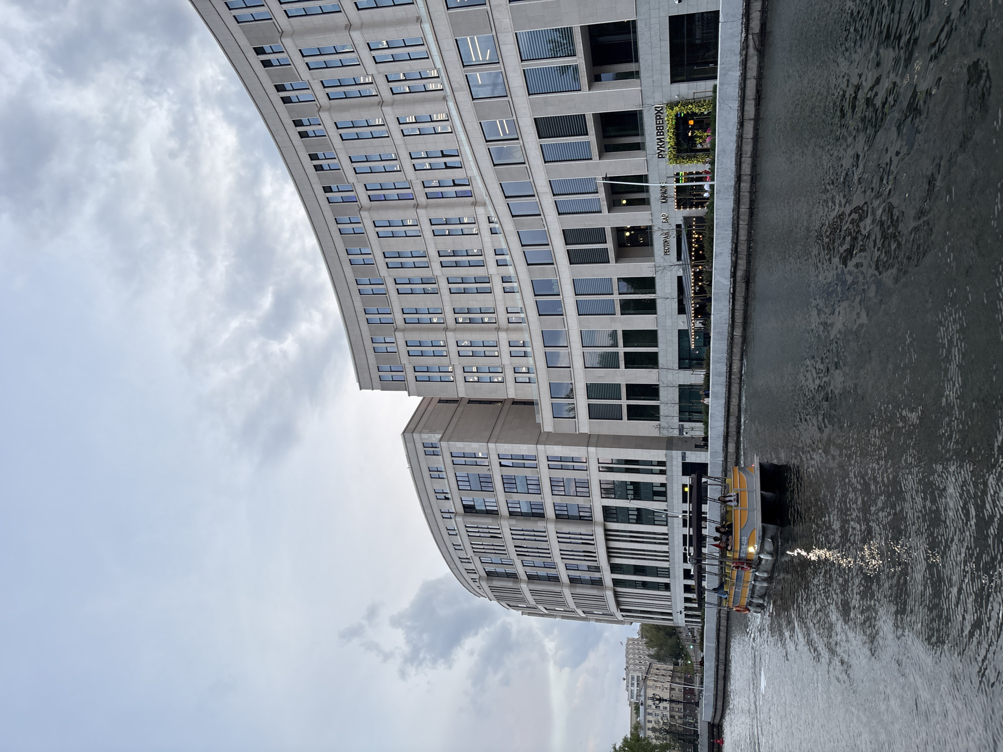 The image shows a view of a modern, curved building with many windows along the Svislach River in Minsk, Belarus. The sky is overcast with grey clouds. A yellow and grey tourist boat is docked along the riverbank, with several people visible onboard. The river is reflecting the light. The building features a restaurant or bar called "Руки ВВерх!" ("Hands Up!"). Other buildings are visible in the distance along the riverbank.