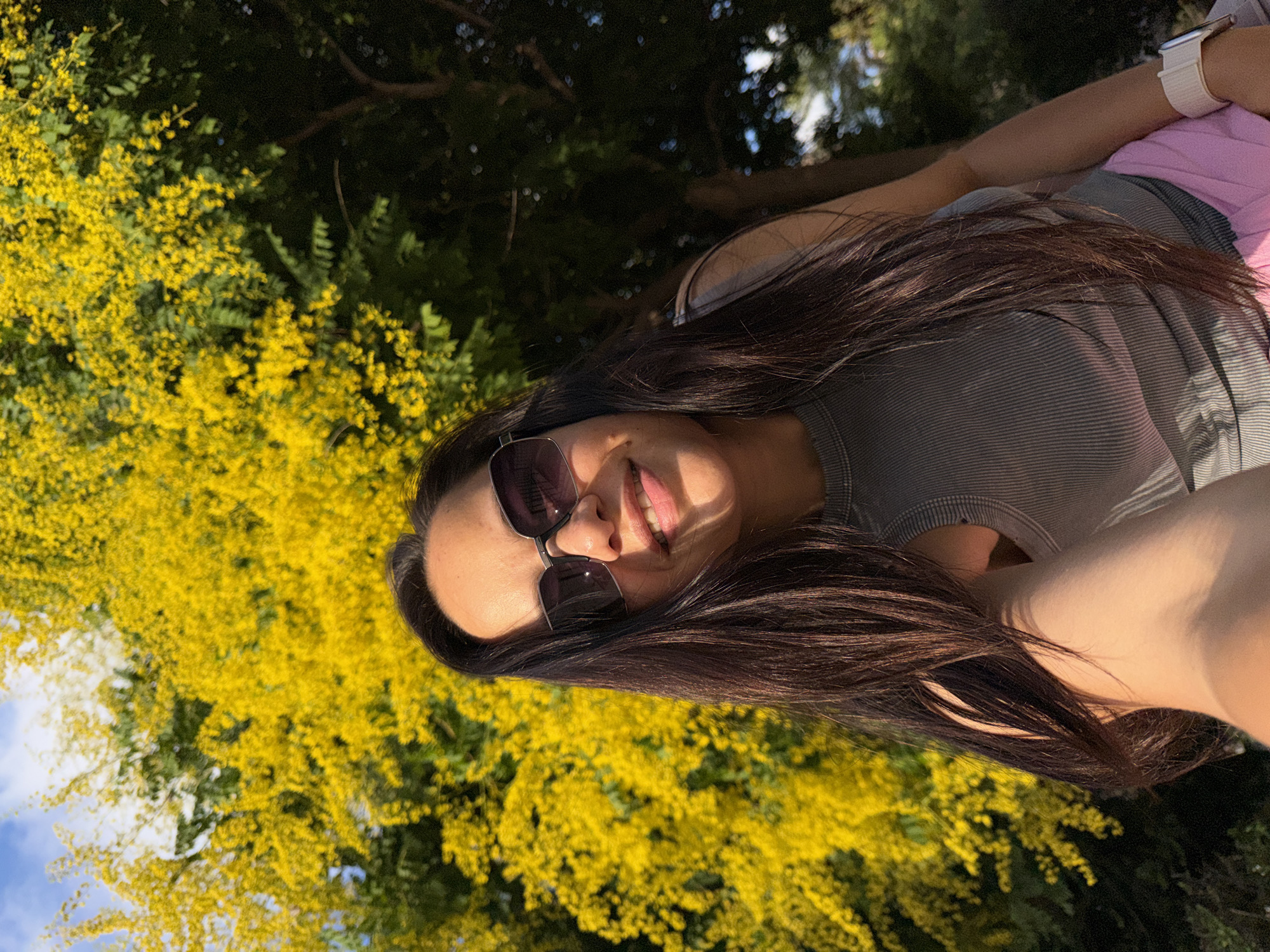 In an outdoor setting, a woman with long, dark hair wears sunglasses and a grey ribbed tank top, along with pink bottoms and a white watch. She holds up a phone, taking a selfie. Behind her, a tree blooms with bright yellow flowers, and lush green foliage, with a sliver of blue sky visible. The scene appears to be in a park or garden.