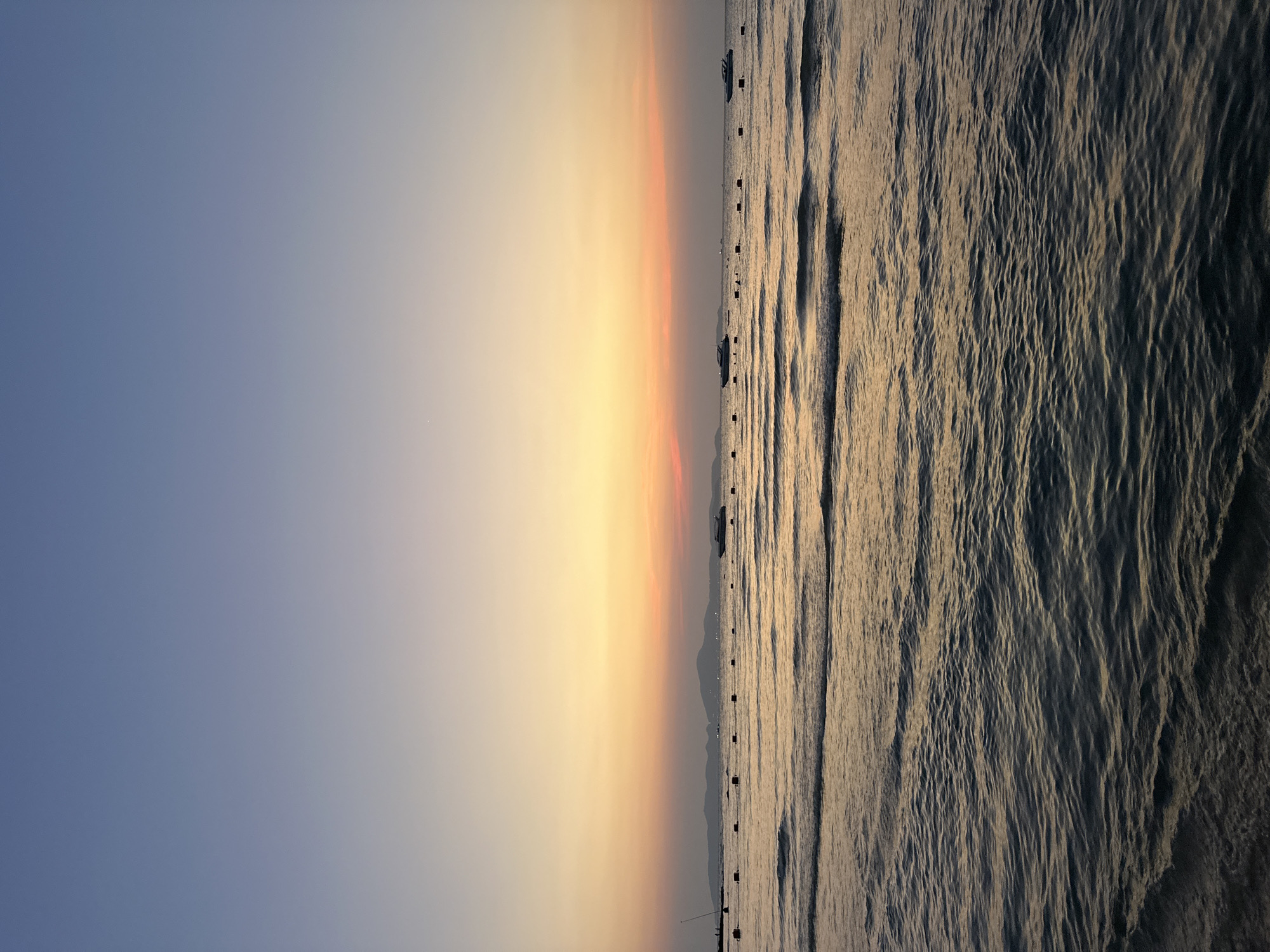 The image depicts a horizontal view of the horizon in Pattaya City, Thailand. To the left, the sky transitions from blue to orange, indicating either sunrise or sunset. The right side of the image showcases the ocean's surface with visible waves. Small boats or buoys are visible on the horizon. The scene suggests a coastal environment with calm waters and atmospheric light.