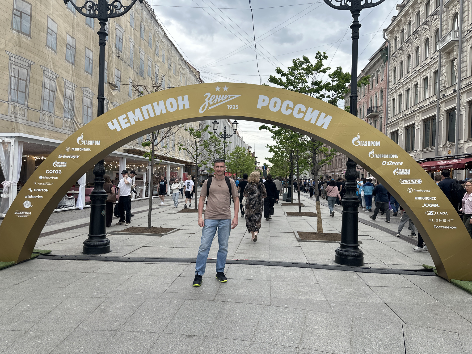 The image shows a man standing in front of a golden archway in Saint Petersburg, Russia. The archway is decorated with the words "Чемпион Зенить РОССИИ" (Champion Zenit Russia) and logos of various Russian companies, including Gazprom, Winline, and Lada. The archway appears to be set up on a pedestrian street with buildings on either side. People are walking around the man and the archway. The sky is overcast. Some buildings are covered by a cloth to conceal ongoing construction.