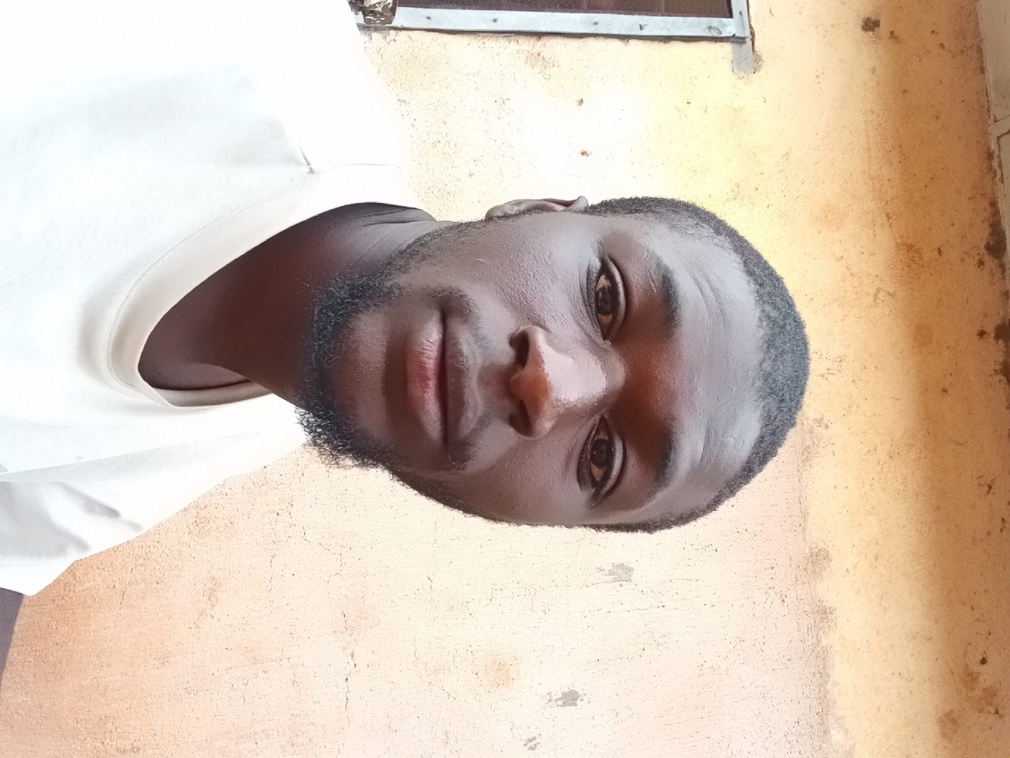 A close-up, head-and-shoulders selfie shows a young Black man with dark skin and short, dark hair. He has a light beard and mustache. He is wearing a white t-shirt. He is looking directly at the camera. The background is a textured, yellow-orange wall with some lighter streaks and imperfections. To the left of the man's head, part of a window frame is visible, with a grey metal frame and a section of a windowpane. The overall impression is of an outdoor setting, possibly a residential or public space in the city of Bukuru, Nigeria, given the wall texture and window details common in the region.
