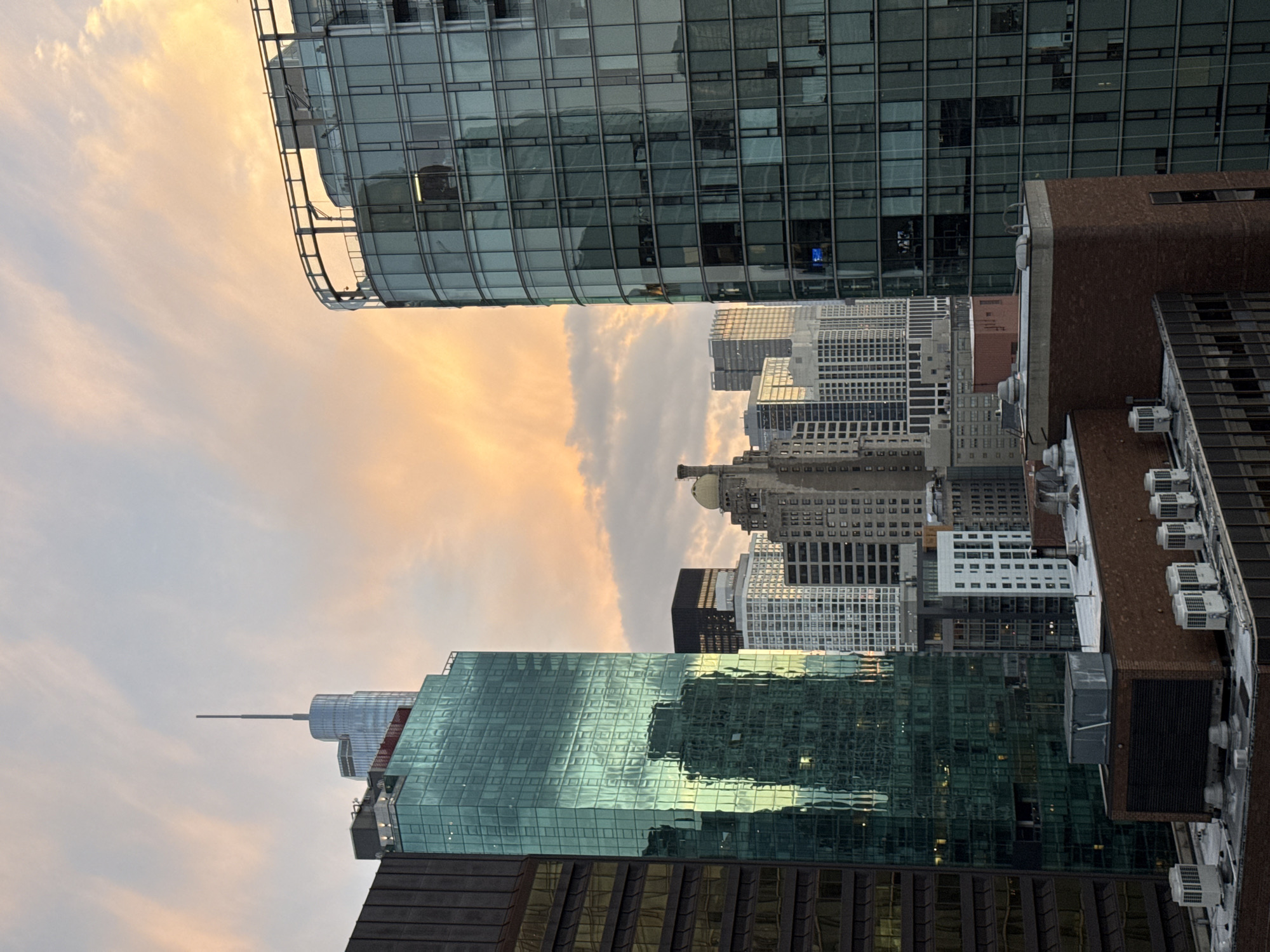 This image captures a cityscape dominated by modern skyscrapers, likely in Chicago, United States of America, given the architectural style and prevalence of glass-clad buildings common in the city. The time of day appears to be either sunrise or sunset, as indicated by the warm, orange and pink hues of the sky and the soft lighting reflecting off the buildings.
In the foreground, a lower brick building with several air conditioning units on its rooftop is visible. Behind it, a tall, dark, rectangular building stands to the left, its facade dark and possibly made of stone or metal. To the right of this dark building is a striking emerald green skyscraper, its glass facade reflecting the sky and other surrounding structures. This green building has a stepped or tiered design.
Further into the background, a variety of other skyscrapers are present. One prominent building has a distinctive rounded glass facade with a visible railing on its upper exterior. Other buildings display more traditional rectangular designs with numerous windows. Some taller structures are capped with spires or antennas. The architectural diversity suggests a central business district or a densely populated urban area.
The sky itself is filled with soft, diffused clouds painted in pastel shades of orange, pink, and light blue, contributing to the serene and visually appealing atmosphere of the scene. There are no people or active events depicted; the image is a static, architectural landscape.