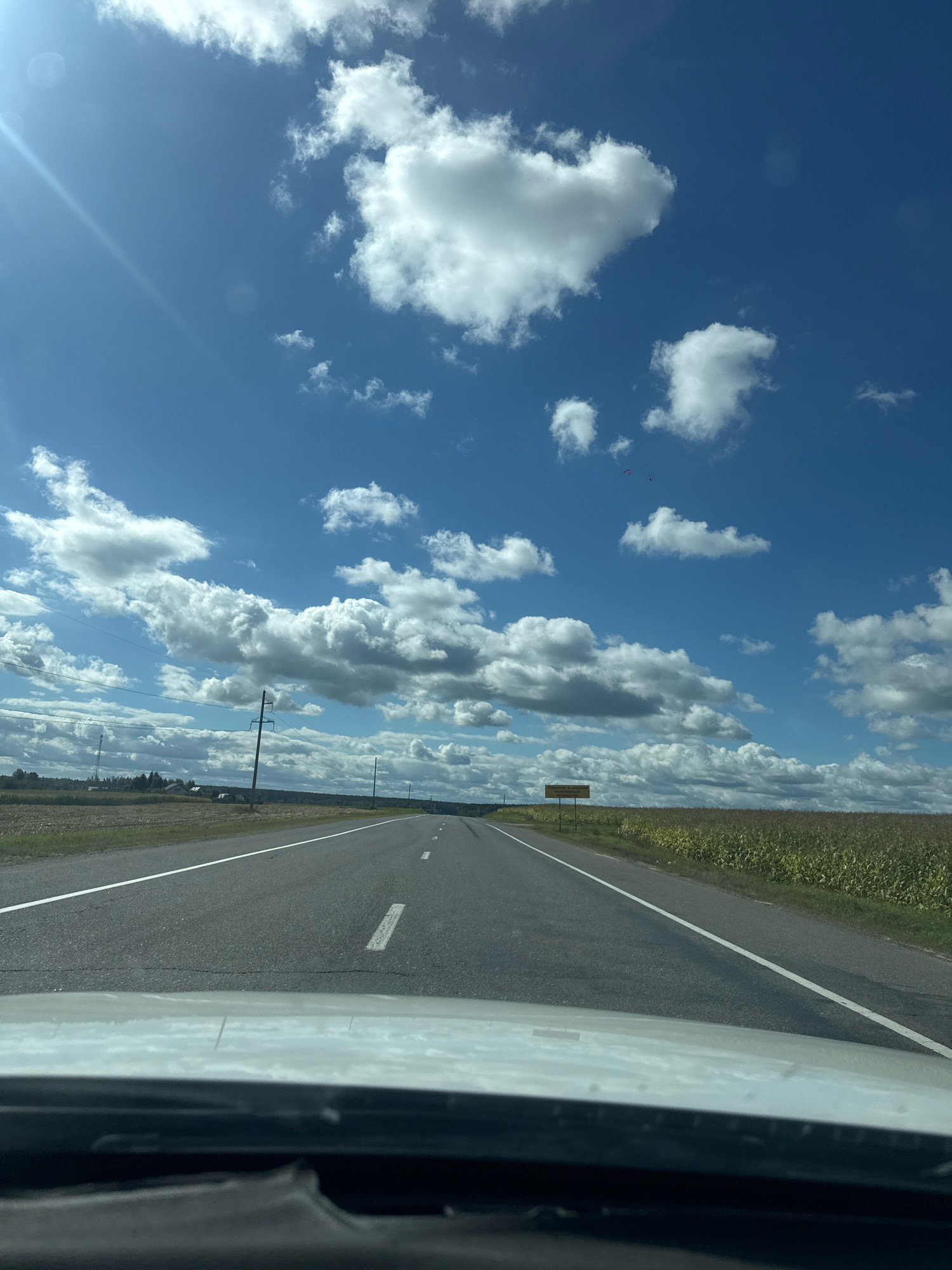 The image depicts a view from inside a vehicle traveling on a paved road. The road has white lane markings and extends into the distance. On the right side of the road, there is a field of corn under a bright blue sky with scattered white clouds. A yellow sign is visible in the distance on the right. On the left side of the road, there are fields and some distant structures, along with utility poles and wires. The sun is shining, creating a lens flare in the upper left portion of the image. The environment suggests a rural or semi-rural setting, possibly in or near the Падгорʼеўскі сельскі Савет, Belarus, given the agricultural landscape and infrastructure.
