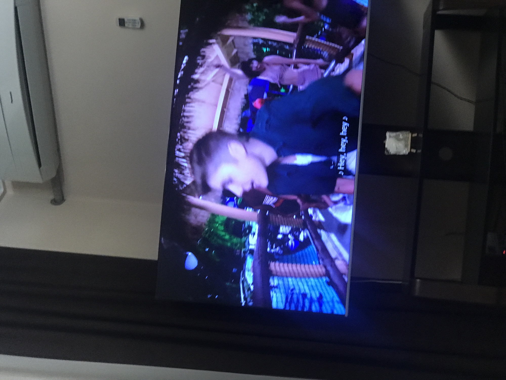 A television displays a scene with a person in the foreground and others in the background, appearing to be in a tropical or outdoor setting. Text overlays the screen. The television is mounted on a dark entertainment center. In the background, a white air conditioning unit is visible on a light-colored wall. The image does not provide any information to determine if this is located in Yerevan, Armenia.