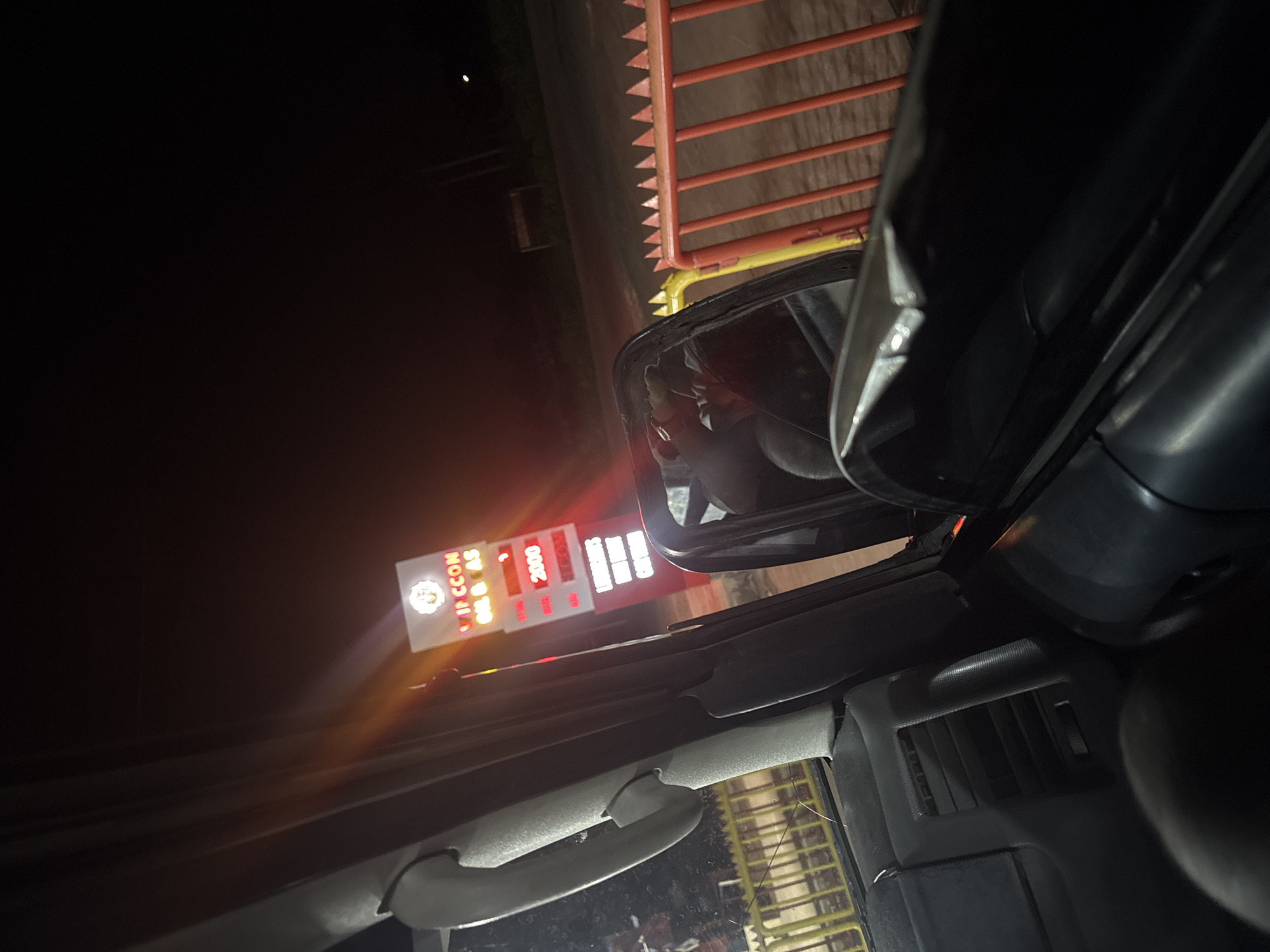 The image is taken from inside a vehicle, likely a car, at night. The driver's side mirror is prominently visible, reflecting a person and a light source. Outside the vehicle, a fuel station sign is illuminated, displaying prices including "2000". A barrier or fence is positioned nearby on the right side of the image. Based on the sign visible in the photo, the location is likely Ozalla, Nigeria.