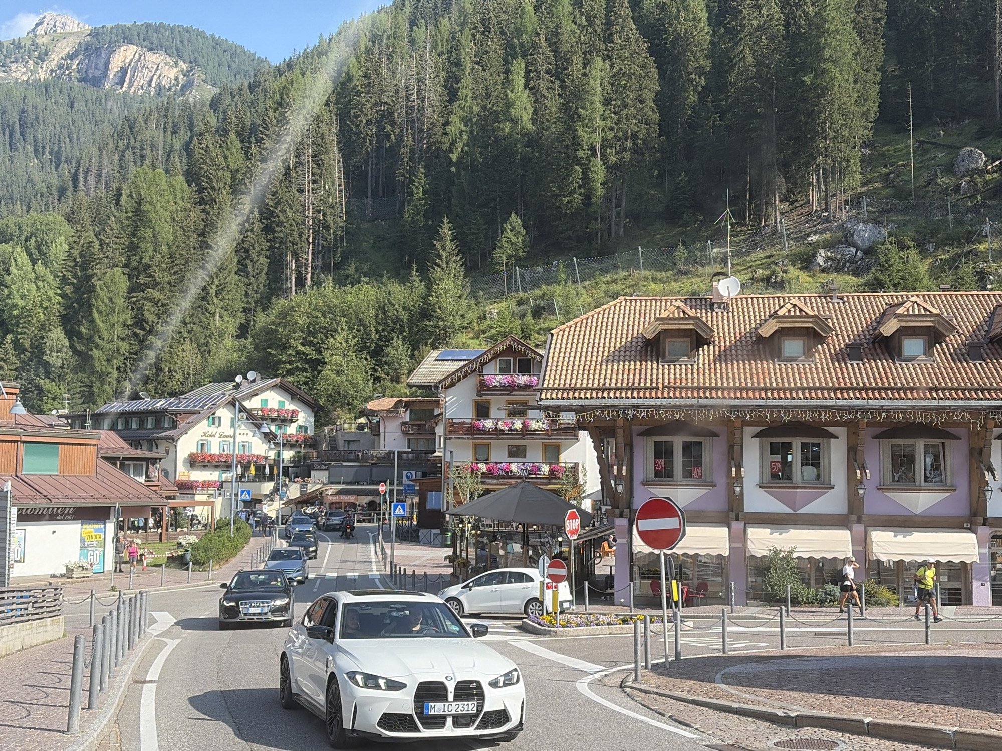 The image depicts a street scene in the city of Canazei, Italy, with a backdrop of a dense pine forest and a rugged mountain peak. In the foreground, a white BMW sedan with license plate "M IC 2312" is driving on a paved road. The car has a black grille and tinted windows. To the right of the BMW, a person in a yellow shirt and dark shorts is walking on the sidewalk. To the left of the BMW, a person in a pink shirt and dark shorts is walking.
Several buildings line the street. One building on the right is a multi-story structure with a tiled roof and large windows, featuring awnings and signage indicating sales with percentages like "-20%" and "-40%". Another building further down the street, labeled "Hotel Loury", has a similar architectural style with balconies adorned with flowers. Across the street, a building with a sign that says "romuren" and "VENDITA" is visible.
Vehicles are also present on the road, including a black Audi and a white hatchback. Traffic signs, including a "STOP" sign, are visible. The environment is characterized by a sunny day with a clear blue sky. A ray of sunlight is piercing through the trees on the mountainside, creating a lens flare effect in the upper left portion of the image. The road is marked with white lines, and bollards are placed along the sidewalk.