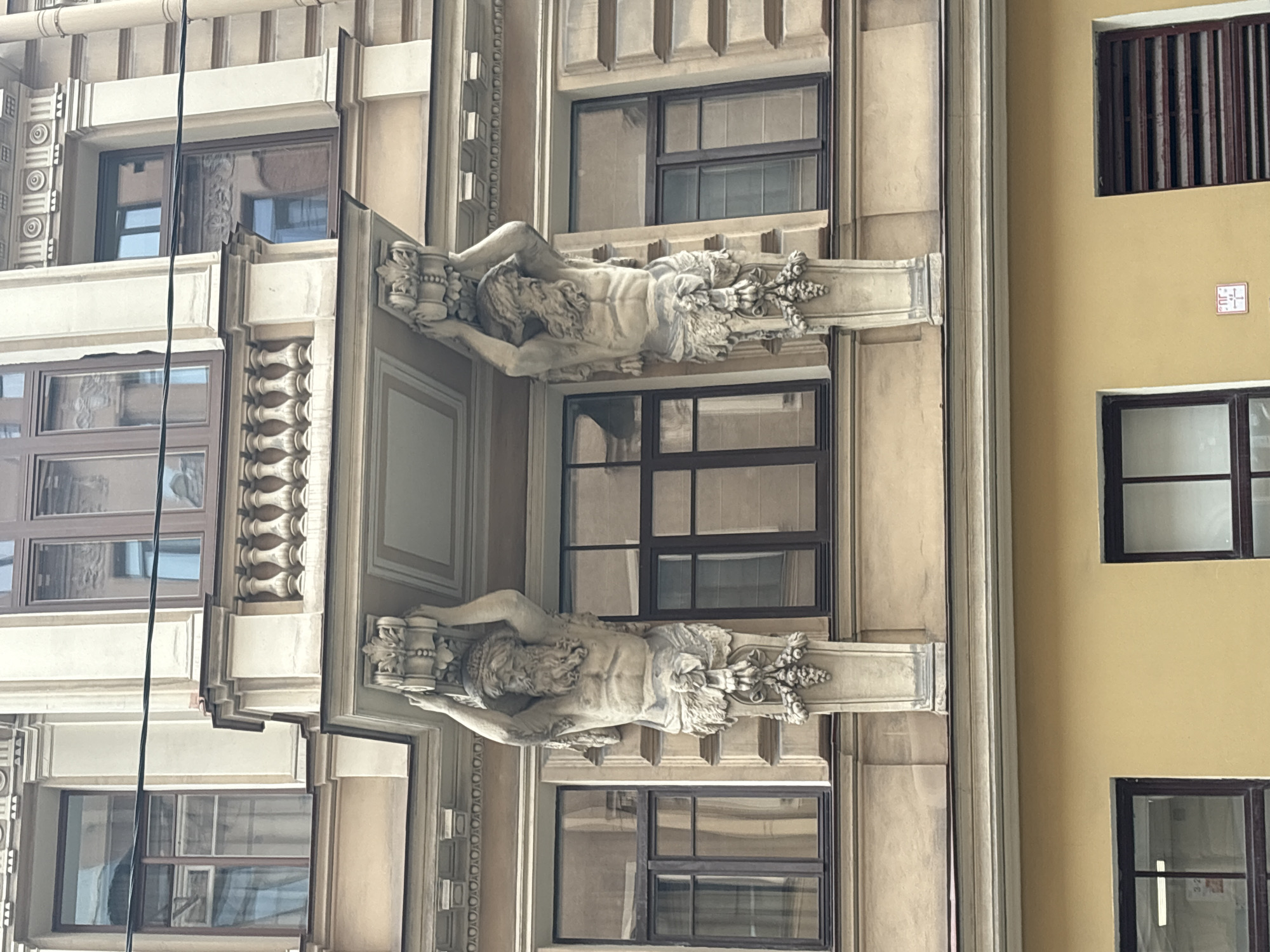 The image shows a building facade in Saint Petersburg, Russia. Two male sculptures adorn the facade, positioned between windows. The sculptures appear to be made of stone or a similar material, and they are lying horizontally with ornate pedestals at both ends. The building has a classical architectural style, with decorative elements and moldings visible. The windows have dark frames. One section of the facade is painted a light yellow color.