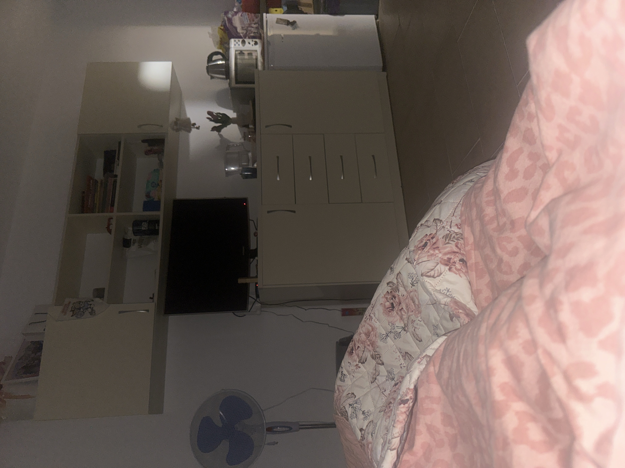 The image depicts a dimly lit room, possibly a bedroom or studio apartment, in Sofia, Bulgaria. In the foreground, a bed is covered with pink and floral patterned bedding. Behind the bed, a floor fan with blue blades stands to the left. A wall-mounted cabinet with open shelves and closed compartments is visible above a long, beige dresser. The dresser has four drawers and two doors. A black television is positioned on top of the dresser. To the right of the dresser, a small white refrigerator is present, with a microwave oven and a kettle on top of it. Various items, including bags and containers, are stored on a shelf above the refrigerator. The overall impression is of a lived-in, somewhat cluttered domestic space.