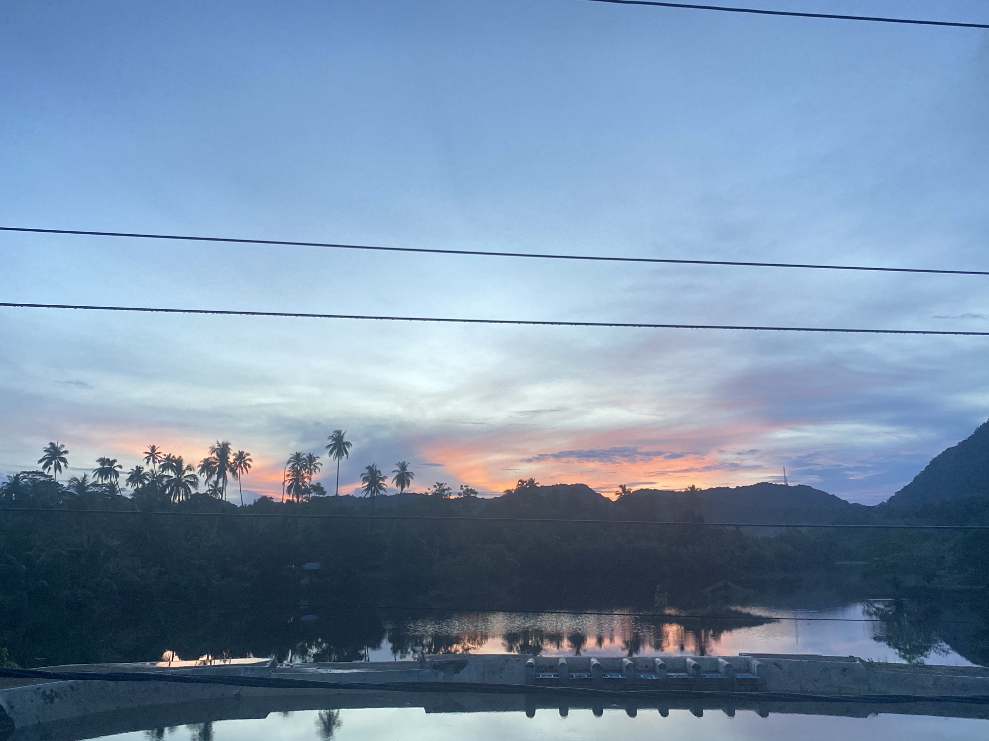 This is a photograph of a tropical landscape at dusk, likely in Lambaro Kueh, Indonesia. The sky is a gradient of blues and purples, with streaks of orange and pink indicating the setting sun behind a silhouetted mountain range. A line of palm trees stands against the colorful sky, their fronds casting dark shadows. Below the trees, a body of water reflects the twilight hues and the shapes of the vegetation. In the foreground, a concrete structure with multiple openings runs horizontally across the bottom of the frame, its surface also reflecting the scene above. Power lines are visible stretching across the upper portion of the image. The overall impression is one of natural beauty and tranquility at the end of the day.