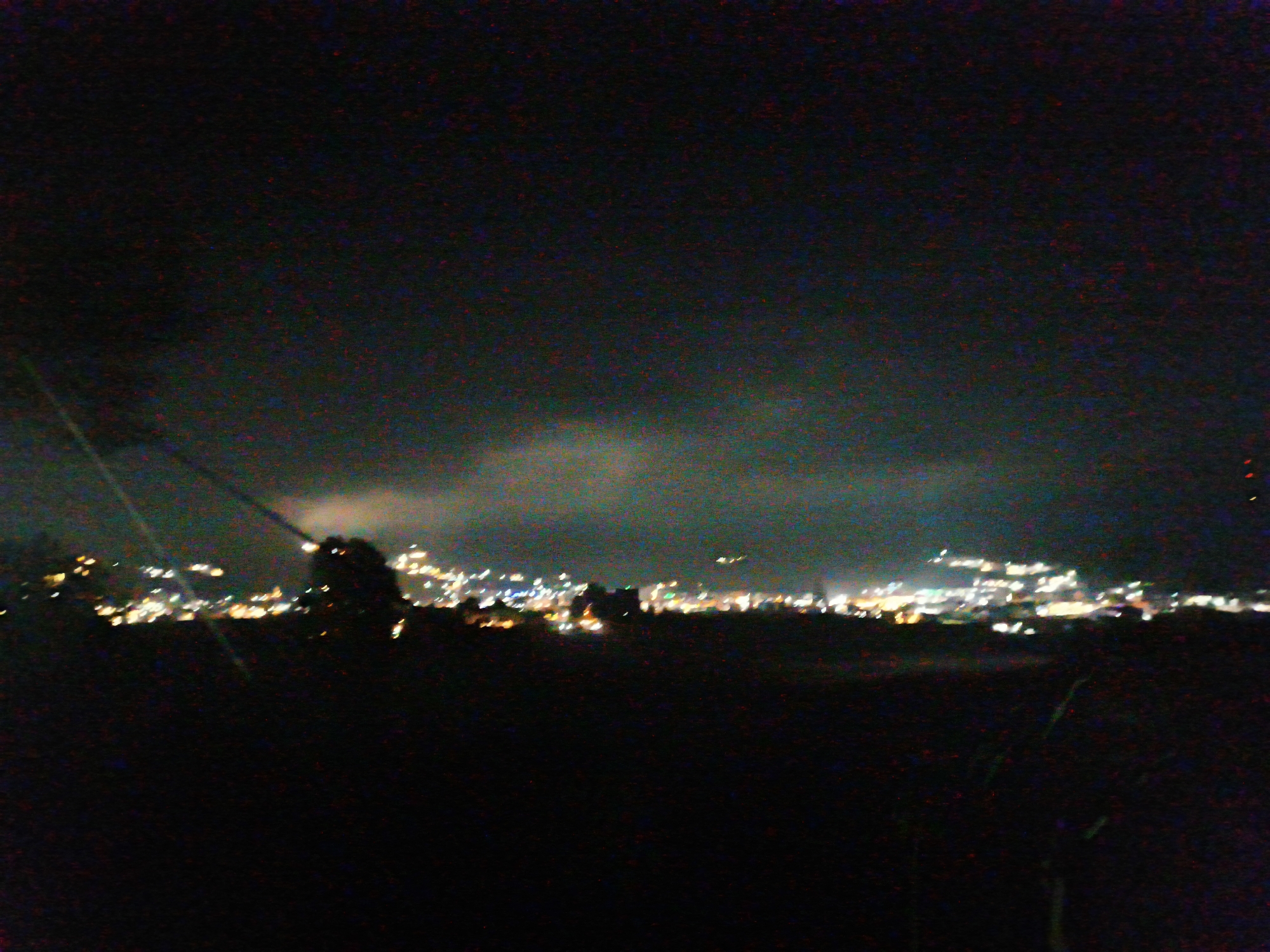 The image depicts a night scene of a city, potentially Dieng Kulon, Indonesia, illuminated by numerous lights. The foreground is dark and out of focus, with the faint outlines of vegetation visible. In the midground, a dense cluster of lights forms the cityscape, suggesting buildings and infrastructure. Some lights appear brighter and larger, possibly indicating significant structures or a brighter area within the city. Above the city lights, the sky is dark and hazy, with faint luminous clouds or atmospheric conditions obscuring the stars. A tall, dark silhouette of a tree or structure is partially visible against the illuminated cityscape. The overall impression is a distant view of a populated area at night, with the darkness and limited clarity of the photograph contributing to a somewhat indistinct and atmospheric representation.