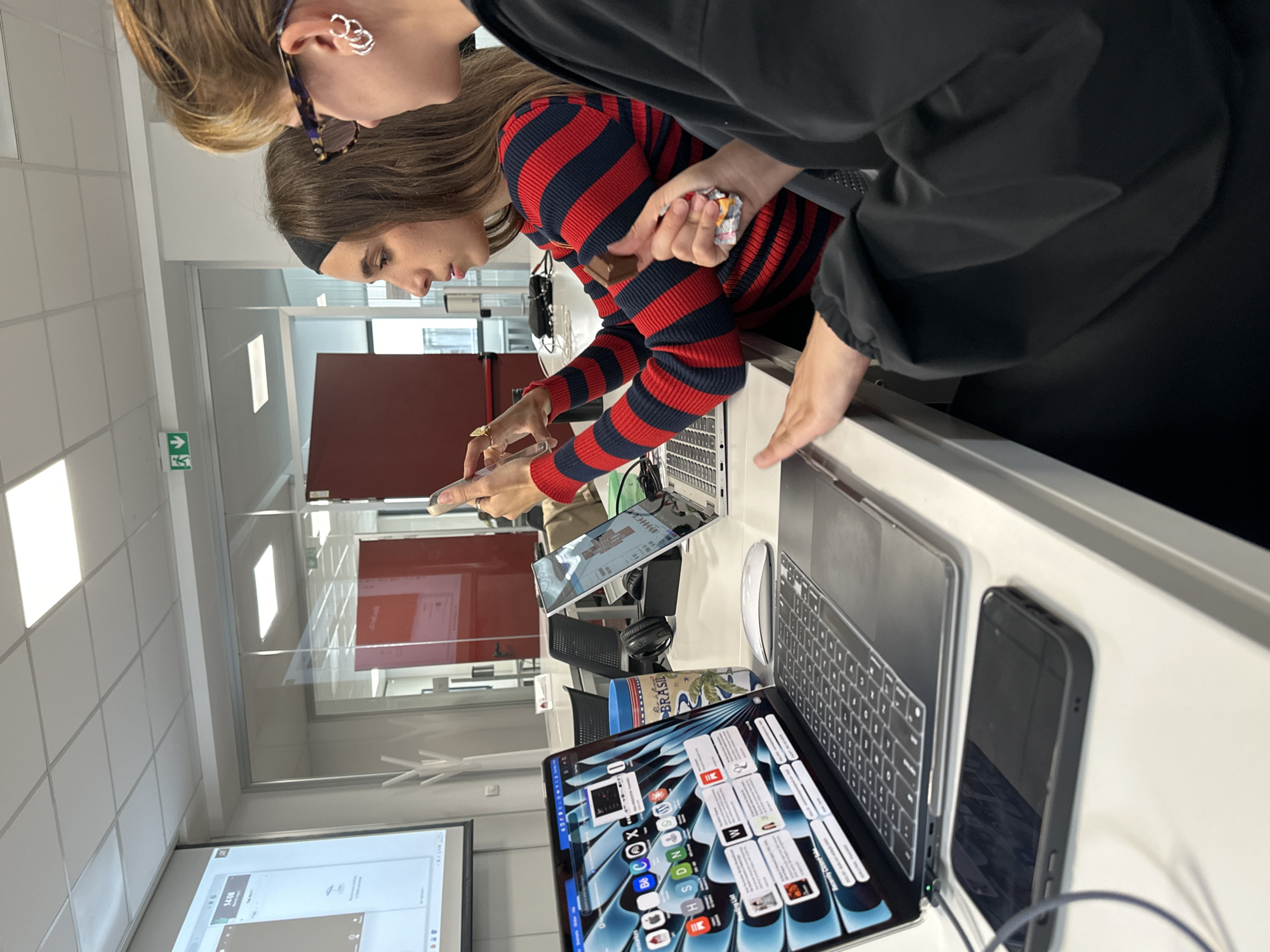 The image shows two women looking at a set of electronic devices on a white surface in what appears to be an office or retail setting. One woman is wearing a red and dark blue striped long-sleeve shirt and is holding a phone and what appears to be a piece of candy or chocolate. The other woman is wearing a black shirt and has sunglasses perched on her head.
The electronic devices include two laptops with their screens visible, two tablets, and a smartphone. One of the laptops displays various icons and a webpage.
The background shows a modern office environment with maroon panels, a ceiling with recessed lighting, and a monitor on the wall displaying a screen. The setting appears to be indoors, with a clean and contemporary aesthetic.
There is no information to confirm that this scene is specifically located in Milan, Italy.
