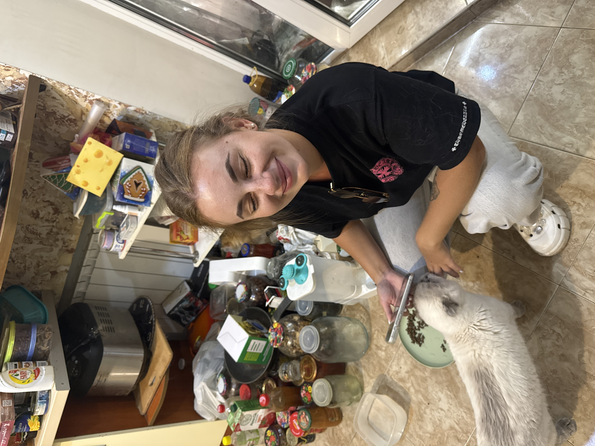 The image depicts an indoor scene, likely a kitchen or pantry area, with a cluttered environment. A young woman is kneeling on the floor, looking down and smiling while holding a smartphone in her left hand. She is wearing a black t-shirt and grey sweatpants, with white crocs on her feet. A light-colored cat with grey markings is interacting with the woman, sniffing at the phone which is placed near a small green plate containing cat food. The background is filled with shelves overflowing with various items, including jars of preserves, food containers, and a bread maker. The floor appears to be tiled. The presence of a bread maker, numerous jars of food, and the general state of clutter suggest a domestic, possibly storage-oriented, space. The user's request for a specific location in Dnipro, Ukraine, cannot be determined from the visual information provided in the image.