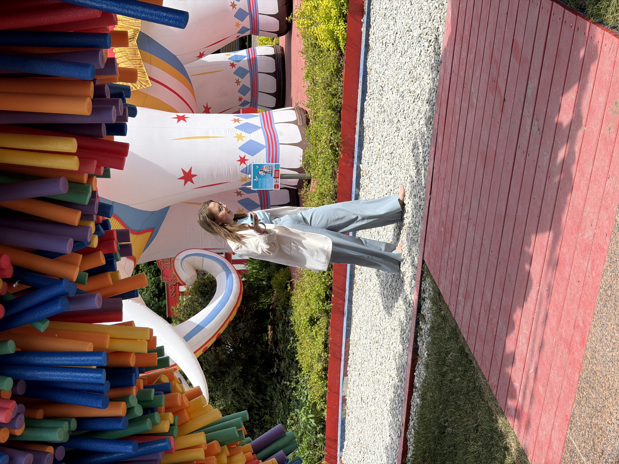 The image depicts a vibrant outdoor scene captured on a bright, sunny day in Moscow, Russia. A young woman, with long light brown hair and wearing glasses, stands in the center foreground. She is dressed in a light-colored blazer over a lighter top, paired with light blue wide-leg trousers, and appears to be barefoot. Her attention is directed upwards and slightly to her right, her mouth slightly open, suggesting she is speaking or reacting to something, with one hand lightly raised in a gesture.
To her left, a dense, colorful installation of foam pool noodles in shades of blue, orange, yellow, red, green, and purple fills a substantial part of the frame, creating a textured wall of color. In the background, behind the woman, are several large, white inflatable structures. These inflatables are adorned with festive red, blue, and yellow patterns, stars, and diamond shapes, resembling giant circus or fairground props. One inflatable features a small blue and white sign displaying an unidentifiable logo and the text "AETO".
Lush green foliage and shrubs are visible behind the inflatables, adding a natural element to the vibrant setting. The ground where the woman stands transitions from light-colored gravel to a red wooden boardwalk on the far right, which is bordered by a strip of green grass and another section of gravel. The clear, sharp shadows indicate strong daylight, suggesting warm and sunny weather. The overall scene conveys a playful and engaging atmosphere, likely at an outdoor event or attraction.