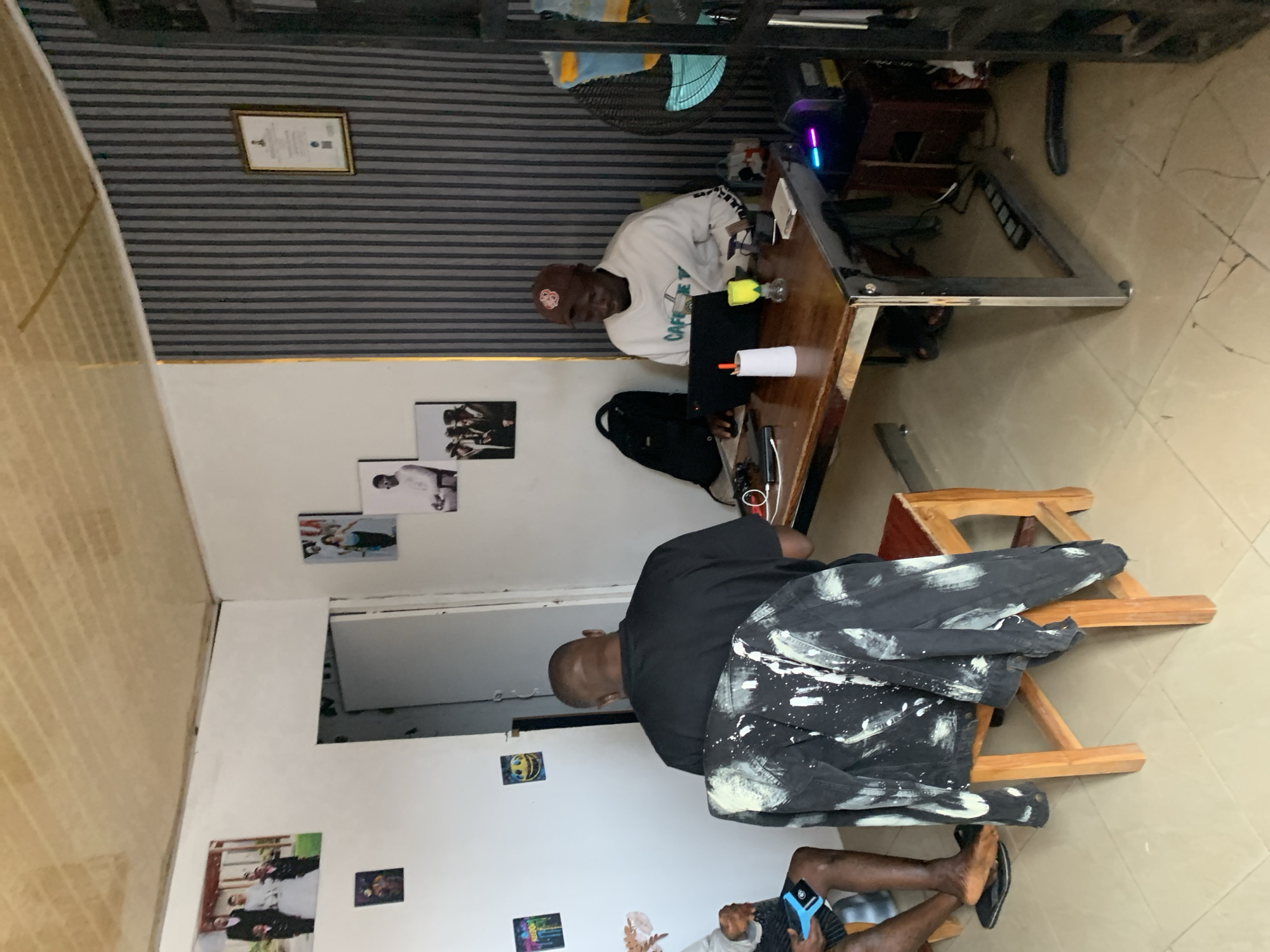 This media file captures an indoor scene in Jos, Nigeria, featuring two adult males in a casual setting. The room has light-colored tiled flooring, white lower walls, and an upper section with gray and black horizontal striped wallpaper or paneling.
In the mid-ground, a man wearing a red baseball cap and a white long-sleeved sweatshirt with the partial text "CAFÉ DE VUE" is seated at a wooden table with a metal frame. He looks towards the viewer with a neutral expression. On the table in front of him are a yellow bottle, a white cylindrical object, and a clear glass item. Behind him, on the striped wall, hangs a framed document, and on the white wall below, several unframed photos are taped. A blue electric fan is mounted on the wall above the table, and a desktop computer with RGB lighting is visible beneath it. A black backpack rests on the floor next to his chair.
In the foreground, with his back mostly to the viewer, sits another man on a wooden stool. He is dressed in a black t-shirt and black pants heavily splattered with white paint. His bare feet, wearing flip-flops, are visible. Next to him, a third person's leg and hand holding a blue-cased phone are partially visible. The white wall behind this man is decorated with more taped photos, including a smiley face and a picture of a wedding couple.
The scene depicts a relaxed environment, suggesting a casual gathering, a shared workspace, or a creative space. The presence of the fan implies a warm climate.
