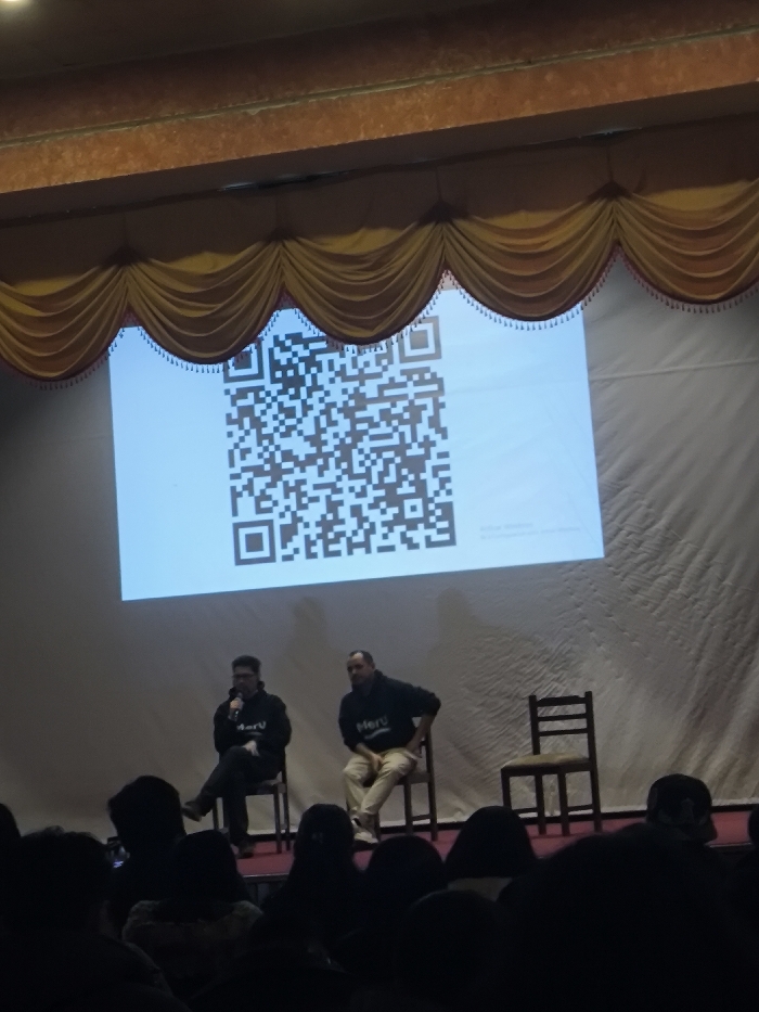 An indoor event is captured on a stage within a large hall in La Paz, Bolivia. A prominent white screen centrally displays a large black and white QR code, with a faint "Activate Windows" watermark visible in its lower right corner. Above the screen, a decorative gold-colored drape with scalloped edges and tassels is visible, beneath an ornate brown architectural molding.
On the stage, two adult males are seated on wooden chairs to the left of the screen's center. Both individuals are wearing dark hoodies with a white "Meru" logo. The male on the left, wearing glasses, is holding a black microphone in his right hand, positioned towards his mouth, indicating he is speaking or about to speak. He is seated with his legs crossed. The male on the right, with short hair and a beard, is seated with his hands resting, attentively observing. An additional empty wooden chair stands to the right of the second male.
In the foreground, the dark silhouettes of numerous audience members are visible, seated in rows, observing the stage. Some audience members appear to be holding up mobile devices. The stage area is brightly lit, contrasting with the shadowed audience section.