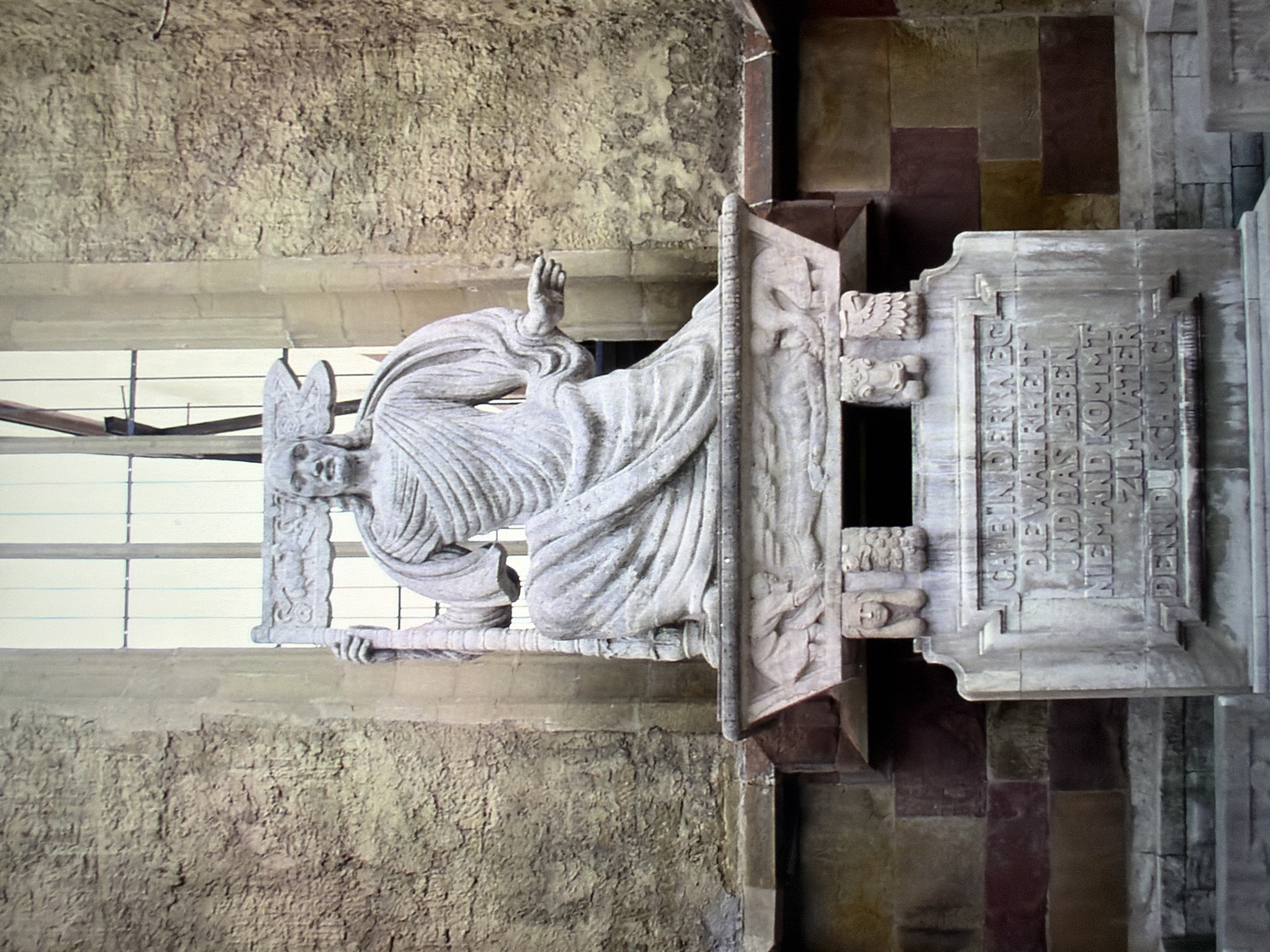 This is a photograph of a stone statue of Jesus Christ, seated and holding a cross. The statue is located outdoors, in front of a large stone building with a tall, narrow window behind it. The statue is highly detailed, with flowing robes and a serene expression on Christ's face. The base of the statue is adorned with carvings, including what appear to be animal heads and figures. The inscription on the base is in German and reads: "Ich bin der Weg und die Wahrheit und das Leben; niemand kommt zum Vater denn durch mich." This translates to "I am the way and the truth and the life; no one comes to the Father except through me."
This statue is located in Kyiv, Ukraine, specifically on the exterior wall of the St. Nicholas Roman Catholic Cathedral. The cathedral is a prominent landmark in Kyiv, known for its neo-Gothic architecture and its history. The statue of Christ is a significant feature of the cathedral's facade, adding to its religious and artistic importance. The stone building behind the statue appears to be part of the cathedral itself, with weathered stone walls and a large, multi-paned window. The overall scene conveys a sense of solemnity and spiritual significance.