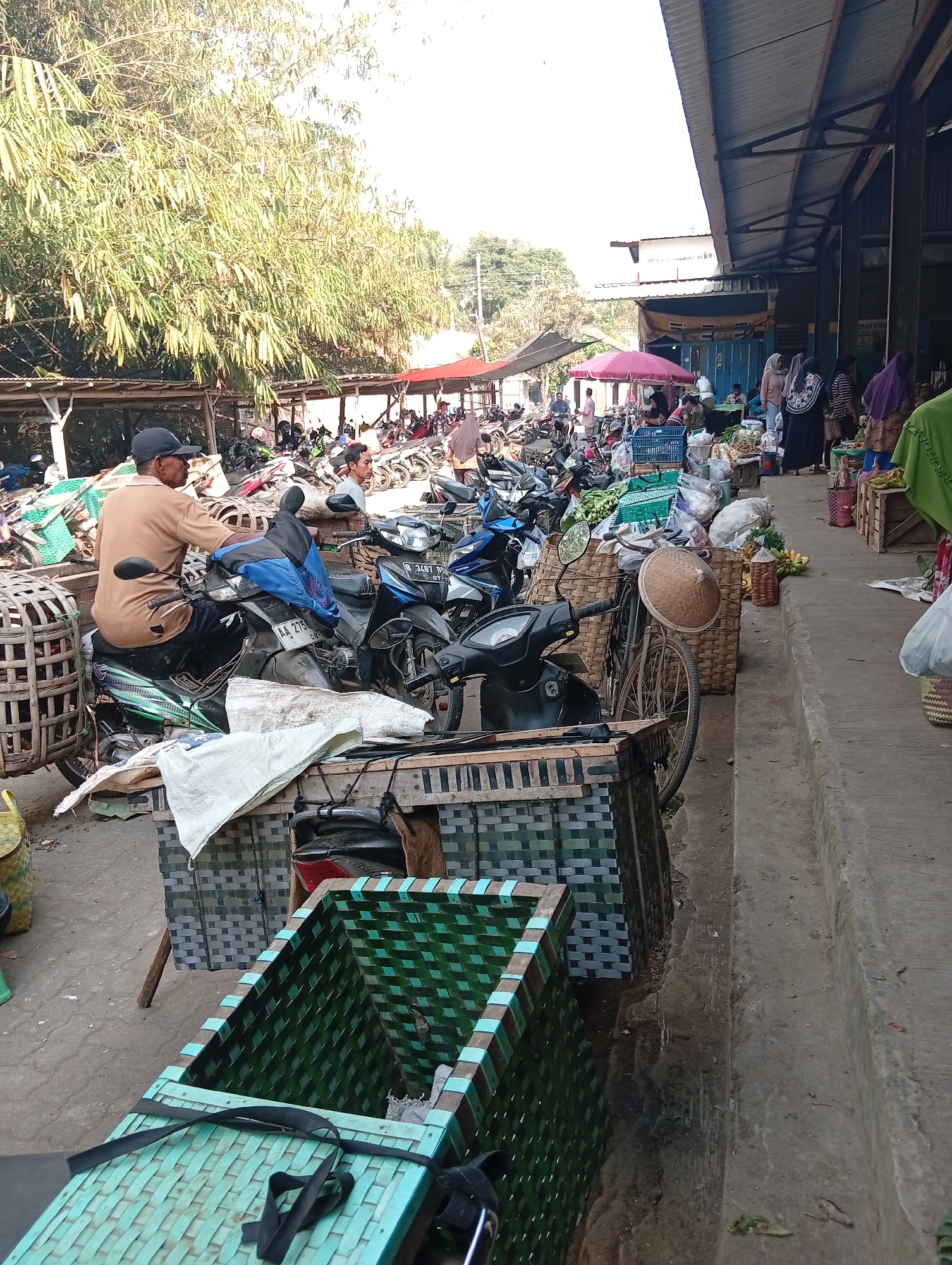 The image depicts a bustling market scene in Plempukankembaran, Indonesia. A man wearing a brown shirt and a black baseball cap sits on a motorcycle, facing away from the camera. To his right, another man in a light-colored shirt is visible. The foreground is dominated by woven baskets and crates, some containing what appear to be produce. Motorcycles are parked in rows, indicating transportation for shoppers and vendors. In the background, a covered market structure with a corrugated roof is visible, where several people, mostly women dressed in hijabs and traditional attire, are seen browsing or working. The overall environment suggests a lively outdoor market, possibly with some covered stalls, and the presence of numerous motorcycles highlights it as a common mode of transport. The lighting suggests it is daytime, with sunlight filtering through the trees and the open market area.