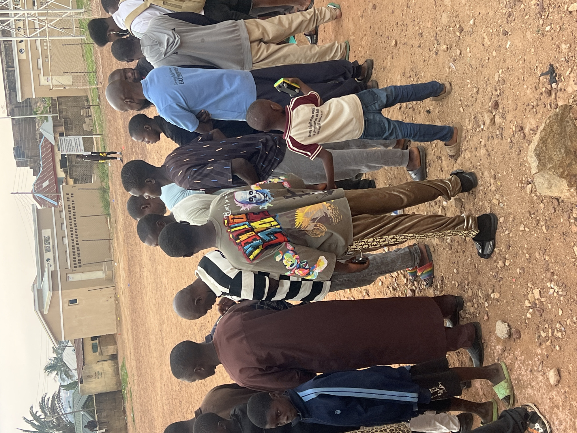 A horizontal eye-level outdoor shot depicts a line of approximately fifteen people, primarily Black males of varying ages, seemingly praying or bowing their heads in unison. The group is standing on a dirt field with small rocks. Most of them are dressed casually, wearing a mix of t-shirts, long-sleeved shirts, trousers, and a jacket. One boy has a toy car in his hand. Buildings are visible in the background, including one with a sign that reads “SOMWA DREAMS PROJECT”. Based on these details, the location is identified as Jos, Nigeria. A person in black and yellow stands in the background near the building.
