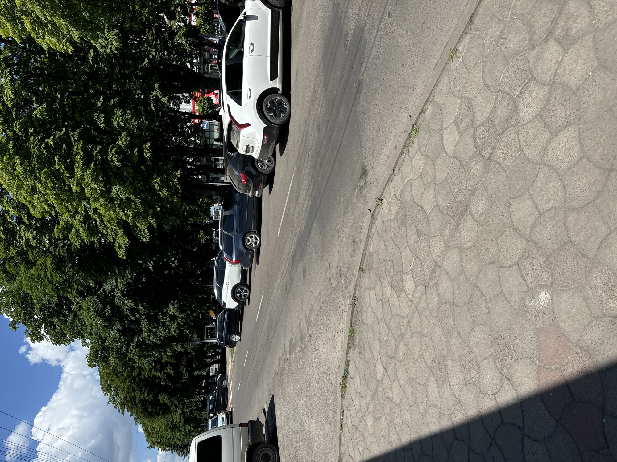 The image shows a row of parked cars along a street in Sarny, Ukraine. The cars are a mix of white and dark colors, and they are parked on the right side of the road, facing away from the camera. The street is paved with asphalt, and there is a sidewalk made of interlocking concrete pavers on the right. Large, leafy trees line the left side of the street, casting shadows on the road and sidewalk. The sky is blue with some fluffy white clouds. The overall impression is of a bright, sunny day in a suburban or semi-urban area. There are no people visible in the image. The activity appears to be simple parking.
Notable details include the distinctive pattern of the interlocking concrete pavers on the sidewalk, which are a common sight in many European countries. The trees are full and green, suggesting it is late spring or summer. The presence of power lines in the lower left corner indicates an urban or suburban setting. There is no visible text in the image.