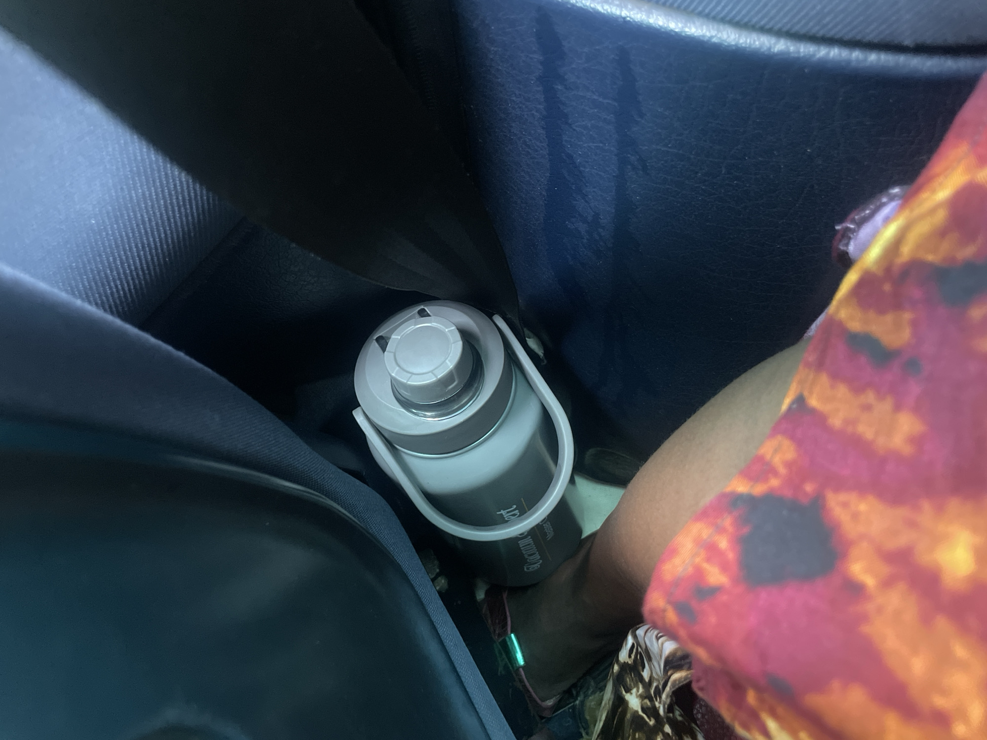 The image shows a person's leg and a water bottle in a car. The person is wearing patterned clothing, possibly a skirt or dress. The water bottle is light grey with a white lid and handle. It appears to be a large, reusable water bottle, possibly a gallon jug. The bottle is placed between the car seat and the person's leg.
The scene appears to be inside a car, with the dark blue interior of the car visible. A seatbelt is also visible, running across the seat. The lighting suggests it is daytime, but the specific time of day and weather conditions are not discernible from the image. The focus is on the water bottle and the person's leg, suggesting the person may be on a journey or outing.
There is some text visible on the water bottle, but it is partially obscured and difficult to read clearly. It appears to be a brand name or some sort of label. The overall impression is a candid shot, possibly taken by the person themselves or by someone else in the car.
The location context provided is Wauro Jabbe, Nigeria. This information, combined with the visual elements, suggests the scene takes place within a vehicle in Nigeria. No specific emotions or actions are strongly conveyed, beyond the practical placement of a water bottle for a person in a car.