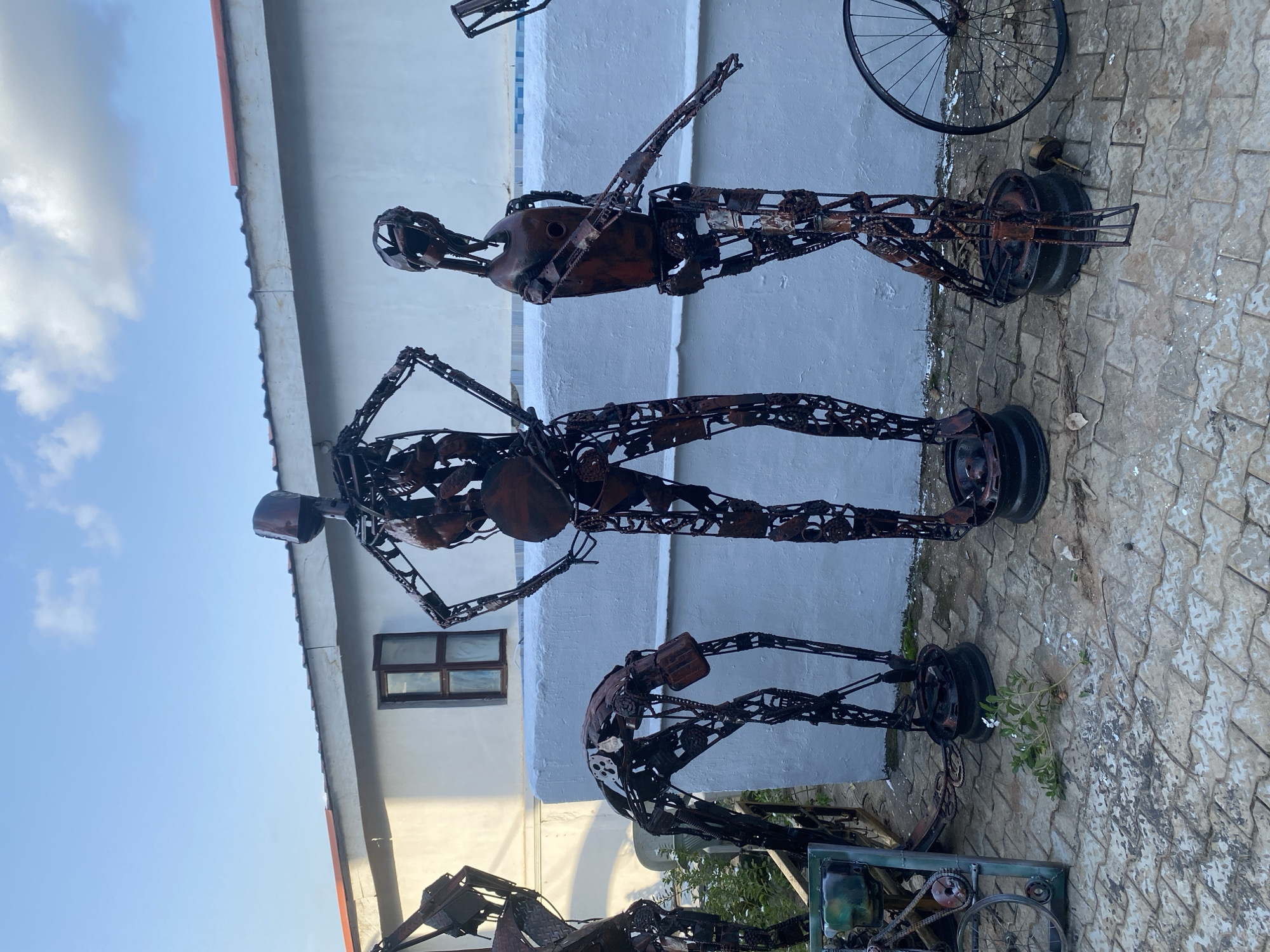 In the city of Unknown, Unknown, a group of five metal sculptures, resembling human figures, are displayed outdoors. The sculptures are made of recycled metal, with visible chains, gears, and other mechanical parts. They are positioned in a row on a paved ground made of interlocking stones. The background features a white building with a red-tiled roof and a single, small, dark-paned window. A white wall with a blue tiled border runs horizontally behind the sculptures. The sky above is a clear blue with a few scattered white clouds. One sculpture is bent over, while another stands upright with hands on its hips. A third figure is positioned as if playing a musical instrument. To the right, another sculpture stands with an arm extended, and a bicycle wheel is placed next to it. A small, green plant with white flowers grows on the pavement in front of the sculptures. The overall scene appears to be an outdoor exhibition or a decorative area.
