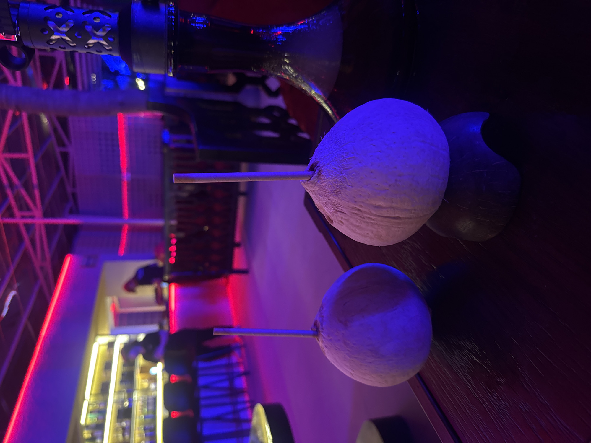 The image captures a scene inside what appears to be a bar or lounge. There are two young coconuts with straws inserted, placed on a dark wooden surface. Behind them, there's a blurred view of the bar area with shelves of bottles and a red and blue neon light installation. The location might be Đà Nẵng, Vietnam, but there are no specific clues in the image to confirm it. The lighting is dim and the atmosphere seems casual and dimly lit.