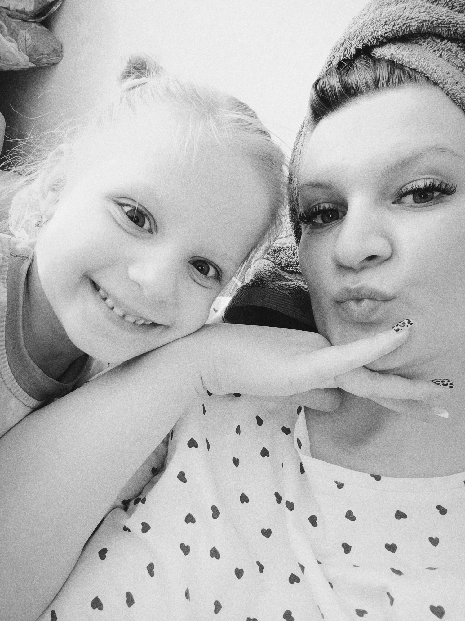 A black and white selfie shows a young girl with blonde hair pulled back in a partial ponytail, smiling with her head resting on her arm, next to a woman with dark hair wrapped in a towel. The woman is making a kissing face. The woman's nails are painted with a leopard print pattern. The woman is wearing a shirt with small heart print. The background is white. The location is not discernible from the image.