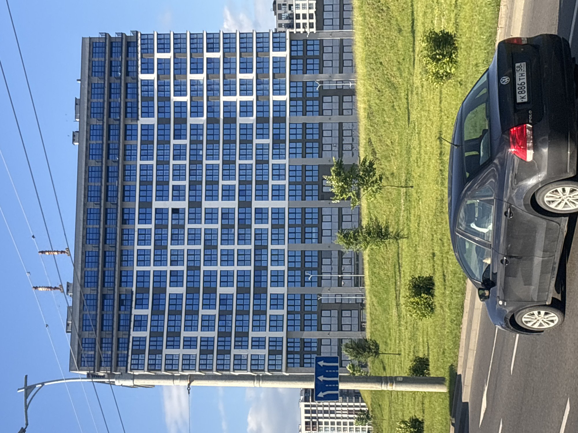 In the vicinity of Сеніцкі сельскі Савет, Belarus, a black Volkswagen Polo sedan is observed driving on a paved road. The car's license plate is visible and reads "K 886TH 58". In the background, a large, modern multi-story building with numerous blue windows and white accents dominates the skyline. The building appears to be an apartment complex or office building. In front of the building, a green, grassy hill slopes upwards. Several small trees and bushes are scattered across the hill. A concrete lamppost with overhead wires stands to the left of the building, and a blue traffic sign with three upward-pointing white arrows is attached to it. The sky is a clear blue with a few wispy clouds. The scene suggests a contemporary urban or suburban environment.