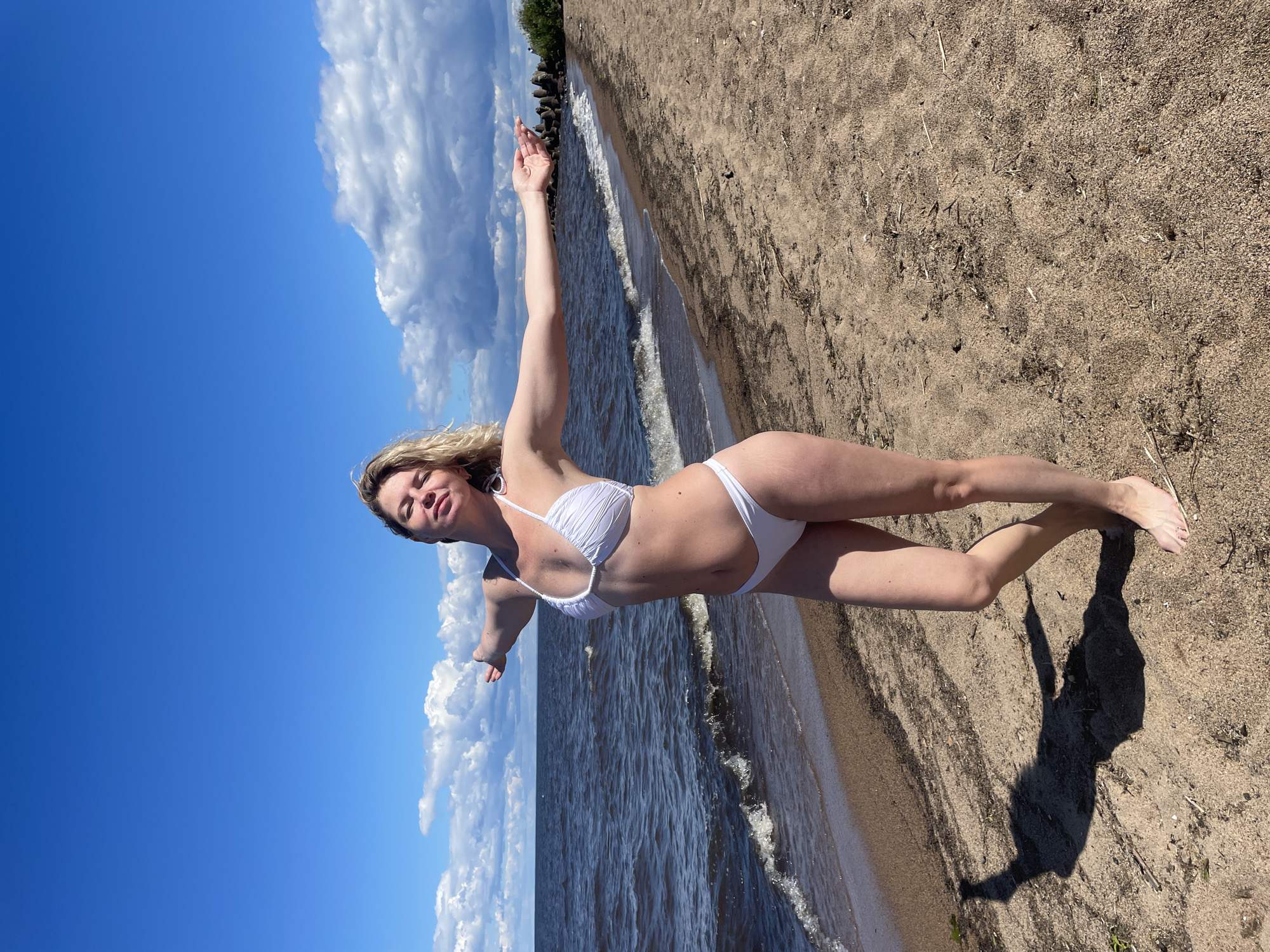 A woman in a white bikini stands on a sandy beach, with her arms outstretched. She is facing the camera, and her eyes are closed, as if enjoying the sun. The ocean is behind her, with gentle waves lapping at the shore. The sky is a clear blue, with a few fluffy clouds. The beach is sparsely populated, with some scattered debris on the sand. In the distance, there are some concrete structures along the coastline, possibly breakwaters. The scene suggests a peaceful and relaxing day at the beach. The location is the city of Belozersk, Russia.