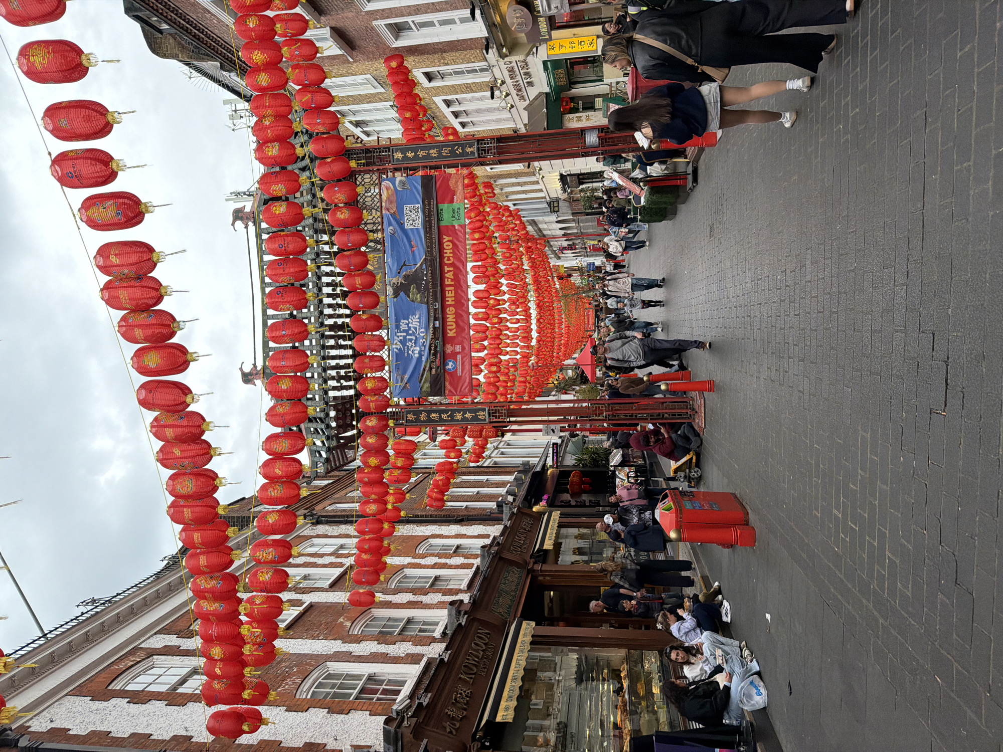 The image depicts a street in London's Chinatown adorned with numerous red Chinese lanterns hanging overhead. Buildings line both sides of the street, with pedestrians strolling along the sidewalk. Several people are sitting on the pavement. Storefronts and signage are visible, including Chinese restaurants and shops. Banners with Chinese characters and English text are suspended above the street.