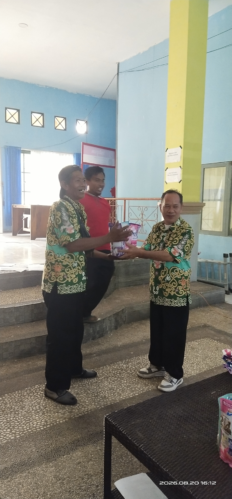 In Tamiang Layang, Indonesia, three men are captured in a moment of interaction within an indoor setting, possibly a school or community hall, indicated by the presence of blue walls, windows, and a yellow column. The man on the left, wearing a green patterned shirt and black pants, smiles broadly as he holds out a package to the man in the center, who is wearing a red shirt and black pants and is also smiling. The man on the right, dressed in a similar green patterned shirt and black pants, reaches out to receive the package. The floor is a textured stone pattern, and stairs are visible in the background. A dark wicker table is in the foreground, with a package resting on it. The timestamp "2026.08.20 16:12" is visible in the lower right corner of the image.