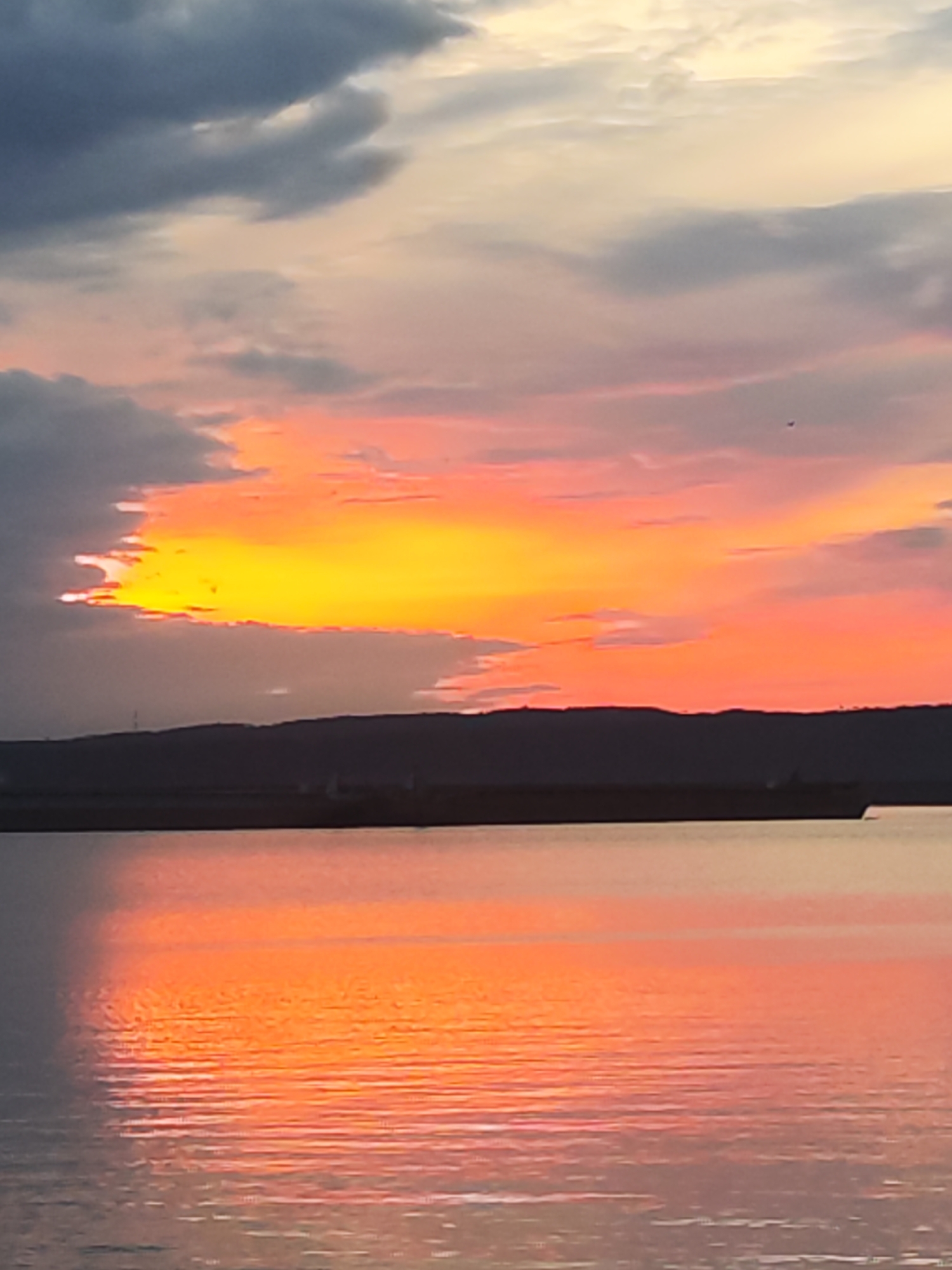 The image captures a breathtaking sunset or sunrise over a vast body of water, likely a river or reservoir in the Chaikovsky, Russia region. The sky is dominated by a dramatic display of colors, with brilliant yellows and oranges concentrated near the center where the sun's light pierces through or illuminates the clouds. These vibrant hues seamlessly blend into softer pinks, purples, and pale grays towards the upper and left portions of the sky, where darker, more voluminous clouds are present.
Across the mid-ground, a dark, undulating silhouette of distant land, possibly a range of hills or a far shore, stretches horizontally, separating the sky from the water. Upon the calm surface of the water, a large, dark, elongated object, appearing as a ship or barge, is visible as a silhouette against the distant land. The water acts as a stunning mirror, perfectly reflecting the fiery orange, red, and pink tones of the sky, with subtle ripples adding texture to the surface. The overall scene conveys a powerful sense of natural beauty and tranquility during the transition of day. No visible text is present in the image.