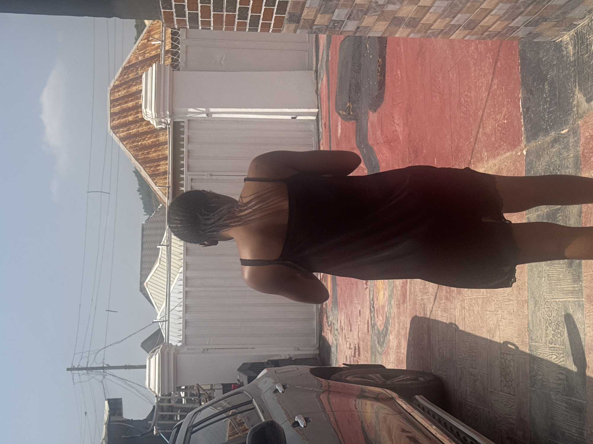 The image shows a person from behind, wearing a dark slip dress. The person has braided hair. They are standing on a patterned ground that is red and black. In the background, there is a white building with a corrugated metal roof and a white gate. There are power lines and a telephone pole visible against a clear blue sky. A car is partially visible in the lower left corner of the image. The scene appears to be outdoors, possibly in a residential area, with elements suggesting a warm climate. The lighting suggests it is daytime.