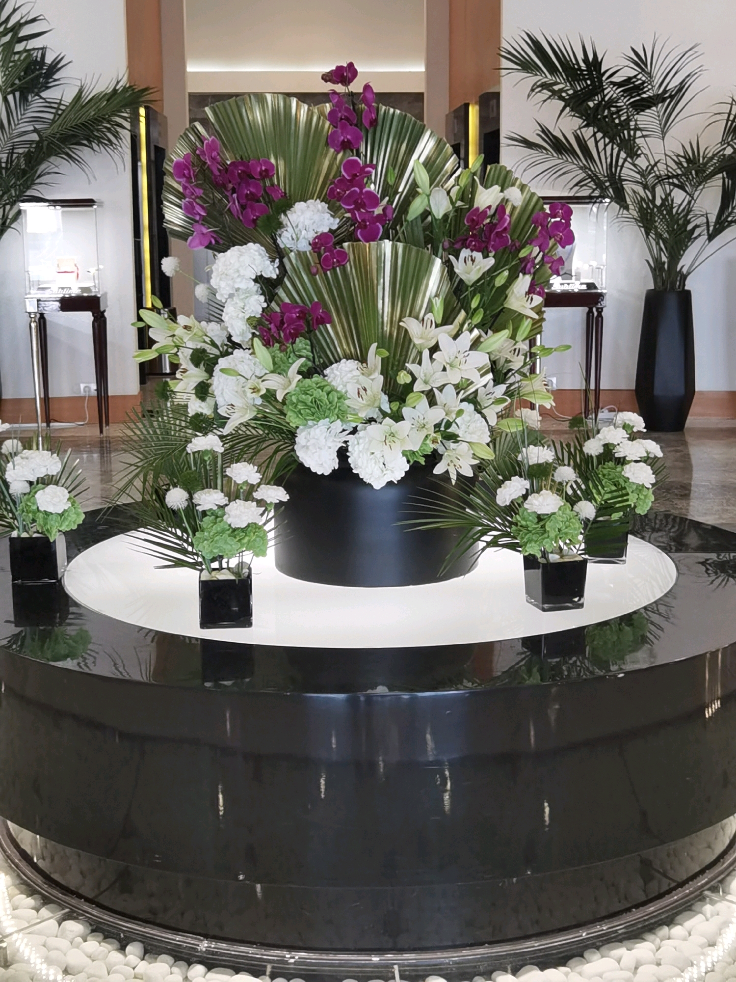 The image shows a large, elaborate floral arrangement displayed in a circular, multi-tiered stand in what appears to be a luxury hotel lobby or similar upscale establishment in Sirius, Russia. The arrangement features a dominant cascade of white lilies and purple orchids, interspersed with lush green foliage and fan-like leaves. Smaller arrangements of white flowers in black square vases are placed on the lower tiers of the stand. Behind the main floral display, glass display cases, possibly for jewelry or watches, are visible, as are tall, potted palm trees in black vases. The floor is a polished dark surface with a circular inlaid pattern. The overall impression is one of opulence and refined decoration, consistent with a high-end commercial space. There are no people visible in the image.