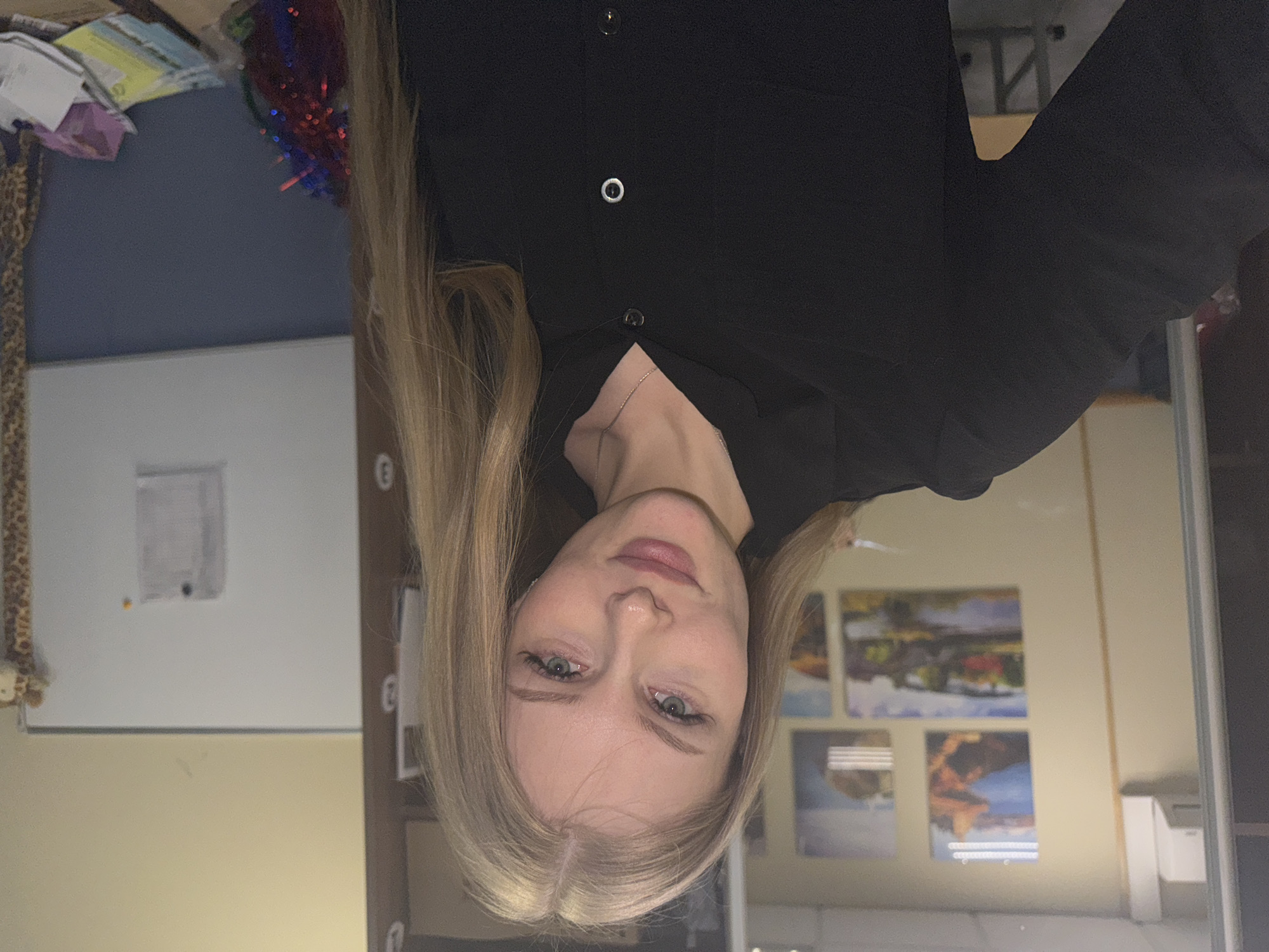 A portrait of a young woman with fair skin, blonde hair, and light eyes is the focal point. She is wearing a black collared shirt with visible buttons. The background includes a whiteboard, a desk with objects, and framed paintings. The location in Perm, Russia is not discernible from the image.