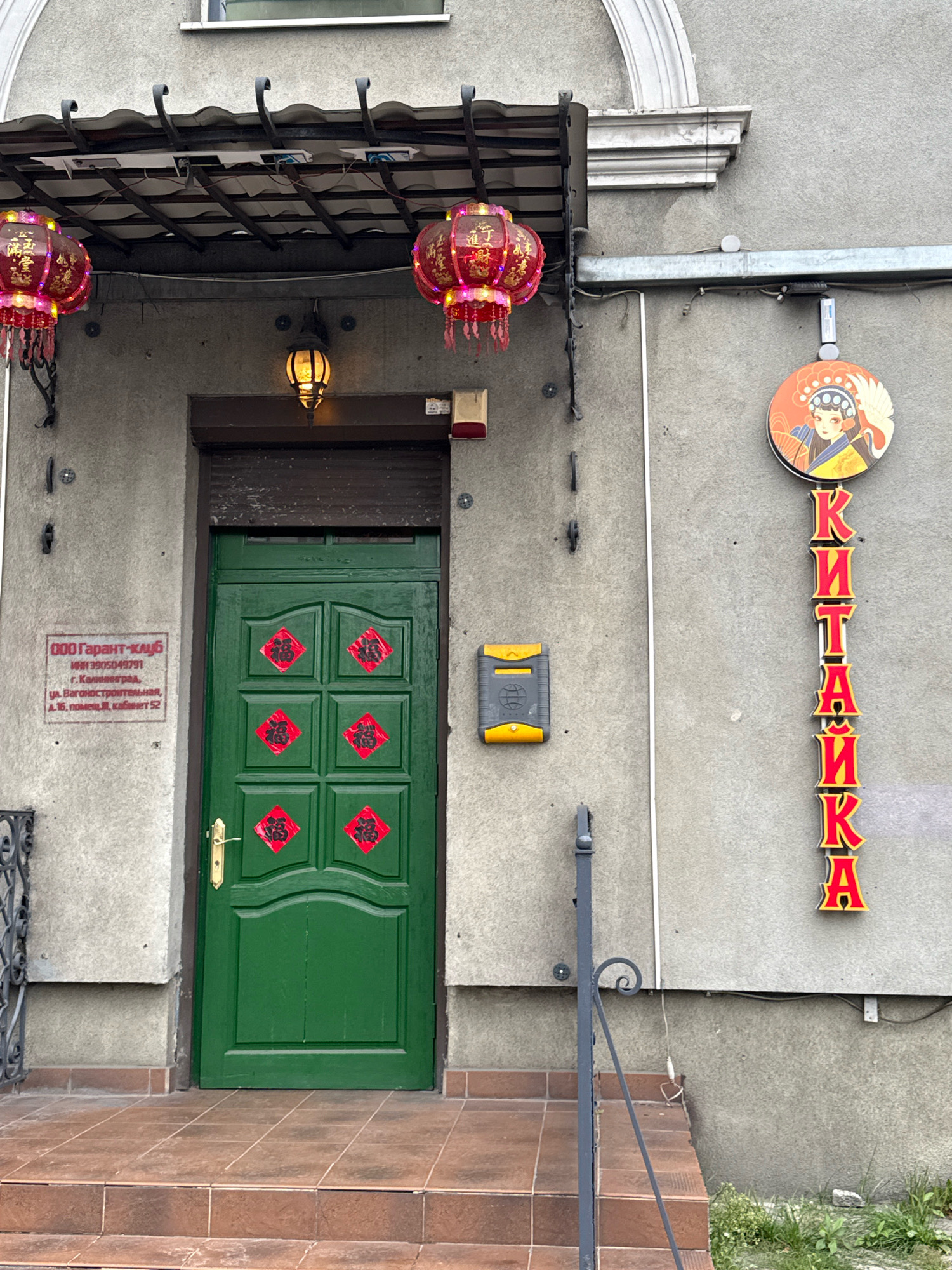 The image shows the entrance to a building, likely a business, in Kaliningrad, Russia. The facade is concrete, with a dark brown, roll-up security door over the main entrance. A green wooden door is visible behind the partially open roll-up door. Red, diamond-shaped decorations with Chinese characters are affixed to the green door, and two red Chinese lanterns hang from the awning above. A circular sign with an image of a woman in traditional Chinese attire and a crane is mounted to the right of the entrance, accompanied by vertical red lettering that spells "КИТАЙКА" (Kitayka). A metal mailbox or intercom unit is mounted on the wall to the left of the door. A business sign in Russian, "ООО Гарант-клуб," is also visible on the wall. The ground in front of the entrance is tiled, with a few steps leading up to the door. A metal railing is present to the left of the steps. The overall impression is of an establishment with a Chinese theme, possibly a restaurant or shop.