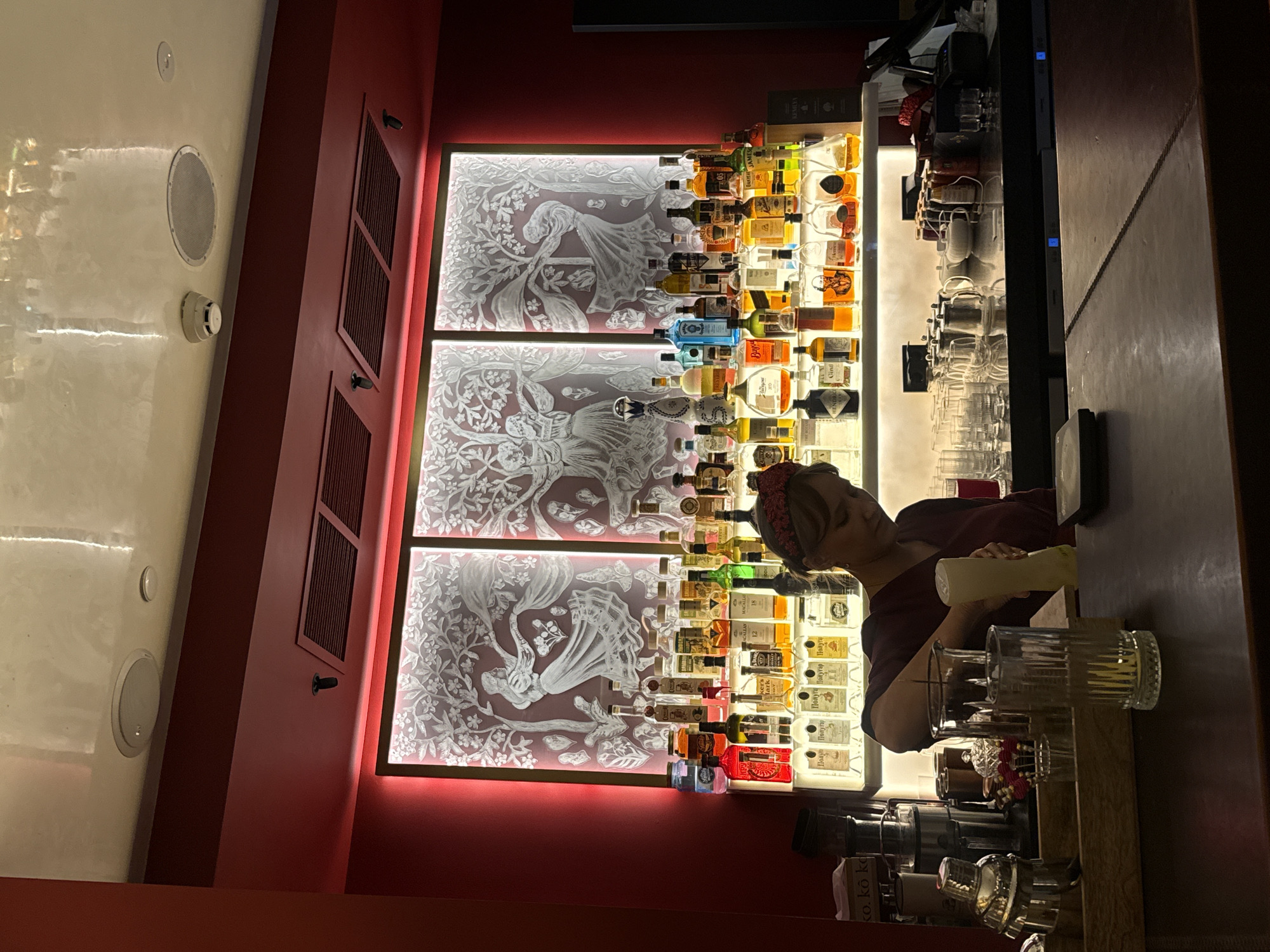 A woman with a red headband is preparing drinks behind a bar. She is wearing a dark red shirt and is pouring liquid from a bottle into a glass. The bar is well-stocked with a variety of liquor bottles displayed on shelves with backlighting. Behind the bottles is a large illuminated glass panel featuring an etched design of people and trees. The bar area appears to be dimly lit, with red walls and a reflective ceiling. Several glasses, bar tools, and a cocktail shaker are visible on the counter. The scene is likely set in a bar or restaurant in Moscow, Russia, given the context provided.