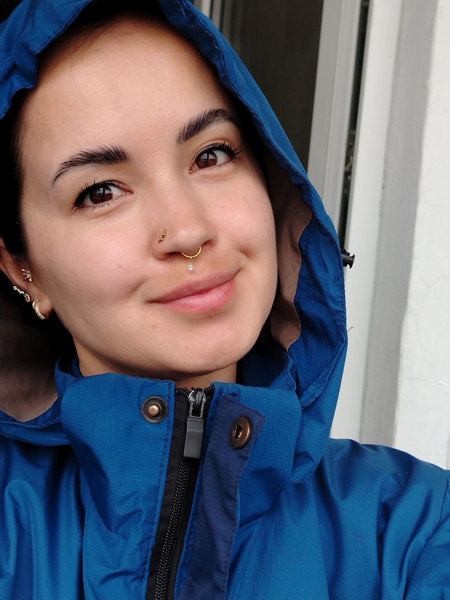 A close-up portrait shows a young woman wearing a blue hooded jacket. She has dark hair, brown eyes, and fair skin. Her facial features include prominent eyebrows and a slightly smiling expression. She has several piercings in her ears and nose, including a nose ring and a lip piercing. The background is indistinct but appears to be an exterior wall or doorway. The location in the city of Noviy, Russia is not evident from the image.