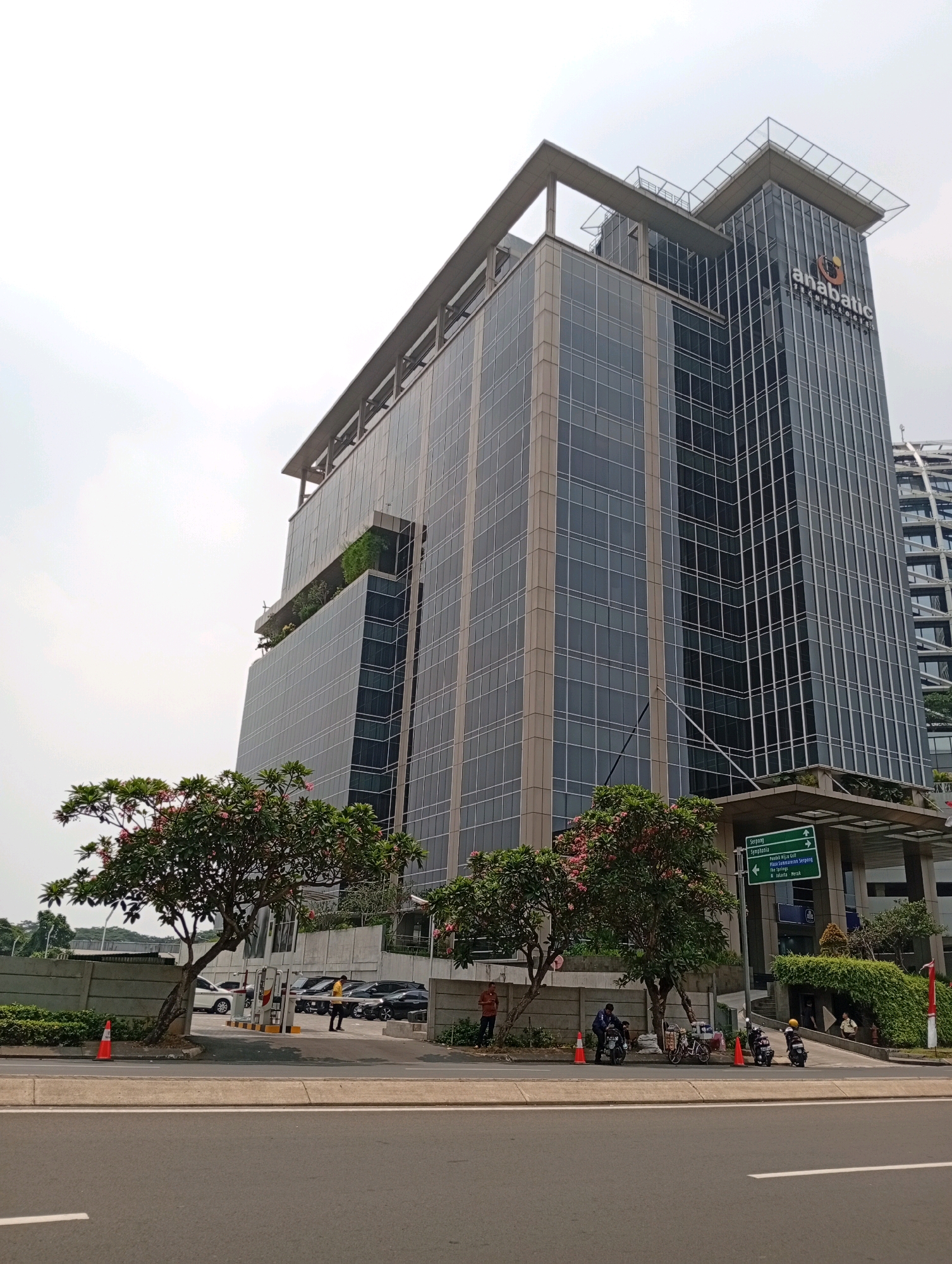 A low-angle, full-shot photograph depicts the Anabatic Technologies building in Curug Sangereng, Indonesia, under an overcast sky. The modern glass and steel skyscraper dominates the scene, its facade reflecting the muted light. The building features a helipad on its rooftop and a logo for "anabatic TECHNOLOGIES" visible near the top.
In the foreground, a street runs parallel to the building, with a concrete median separating it from the sidewalk and the building's entrance area. Several vehicles are parked or driving on the street and in the parking lot. Trees with pink blossoms flank the entrance to the building, providing a splash of color against the urban backdrop.
On the sidewalk near the building entrance, a few people are visible, some standing and some appearing to interact with motorcycles and bicycles. Orange traffic cones are placed along the edge of the road and sidewalk. A green directional signpost stands near the building's entrance, indicating directions to various locations, including "Serpong," "Symphonia," and "Plaza Senayan."
The environment suggests a typical urban setting with a mix of modern architecture and greenery. The overall impression is one of a bustling commercial district.