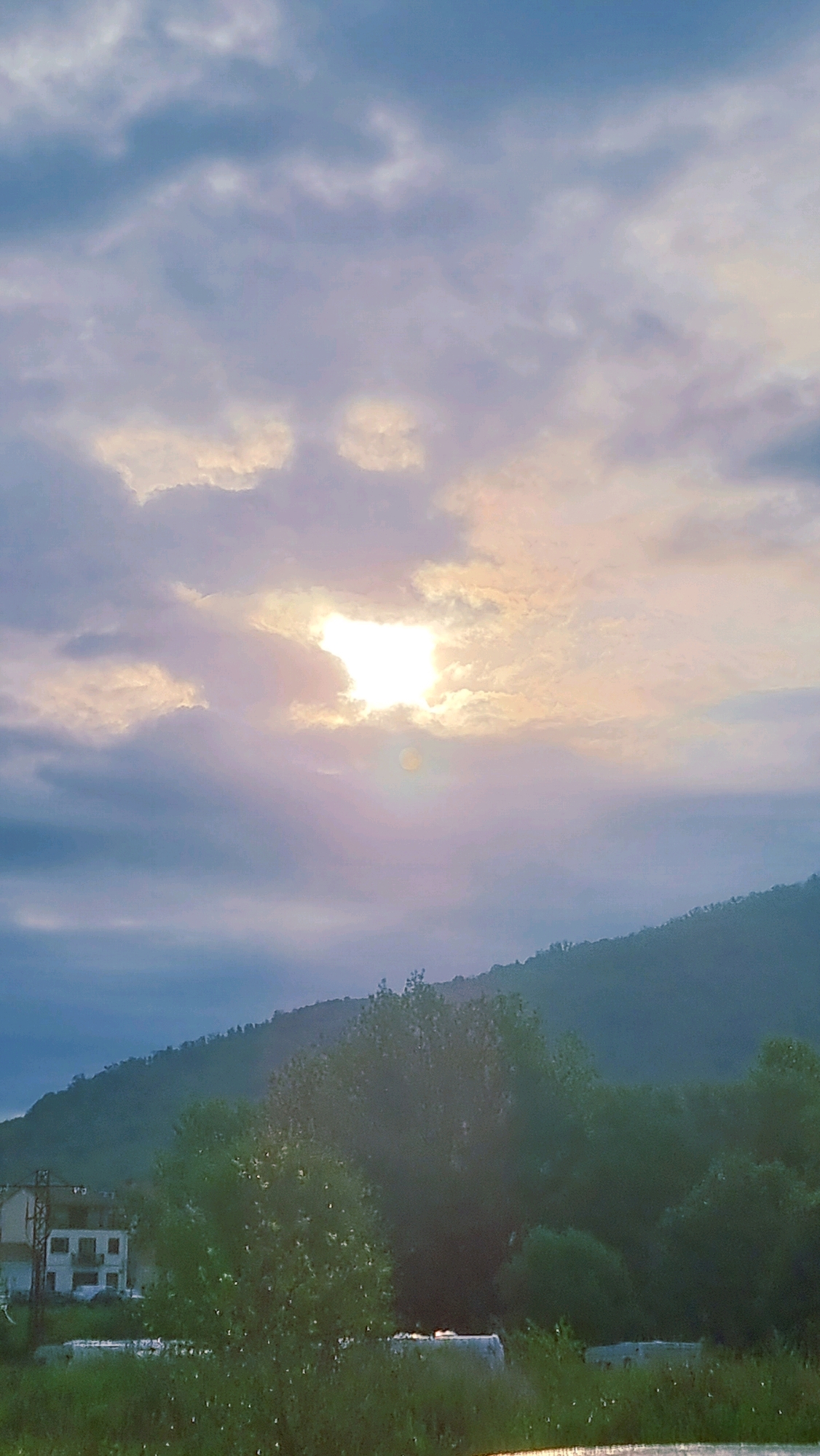 The image depicts a landscape scene with a dramatic sky and a hint of human habitation. The sky is filled with a mix of light and dark clouds, with the sun peeking through a gap, casting rays of light downwards. The lower portion of the image shows a green hillside covered in trees. In the foreground, there is a small white building with a red roof and a balcony, suggesting a rural or semi-rural setting. The overall atmosphere is serene, with the natural elements dominating the composition. The location appears to be in the municipality of Sant Joan les Fonts, Spain, known for its natural beauty and historical sites.