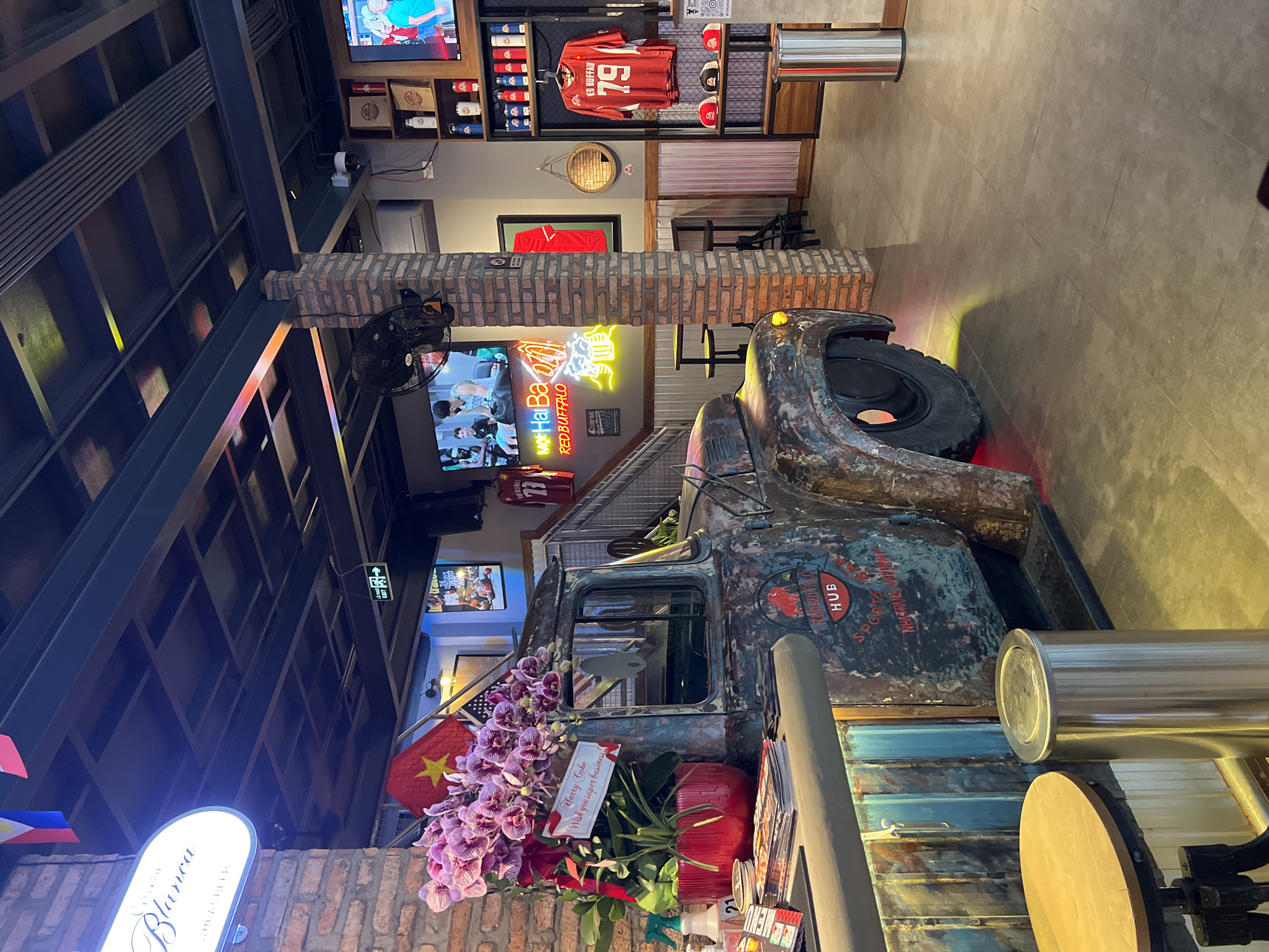 This is an indoor scene, likely a restaurant or bar in Bắc Nha Trang, Vietnam. The decor includes a weathered, vintage truck front end that has been repurposed as part of the interior design. The truck's paint is faded and peeling, but the "Red Buffalo Hub Sports" logo is still visible. There are Vietnamese and possibly other flags (like the Philippines) displayed alongside the truck. Fresh flowers, including orchids and roses, are arranged around the truck, accompanied by a card addressed to "Harry Cohn" wishing him "super business." Neon signs and television screens showing sports events or other programming are visible in the background. Sports memorabilia, including a jersey and hats, are displayed on shelves. The restaurant features exposed brick elements and a contemporary industrial design with metal framing and open shelving.